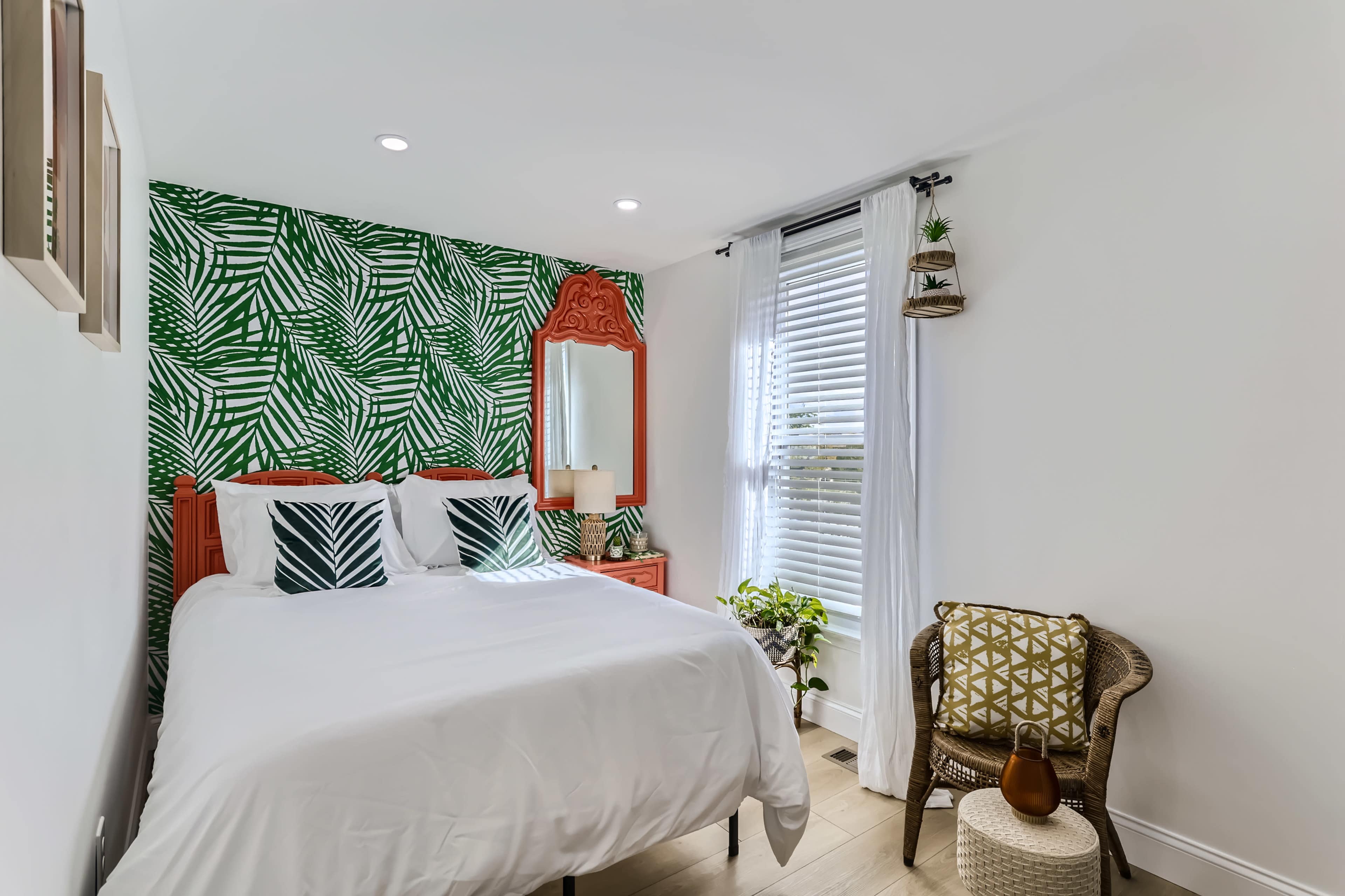 A compact bedroom featuring a bed with white linens, a green tropical leaf-patterned accent wall, and a small sitting area with a chair and decorative pillows.