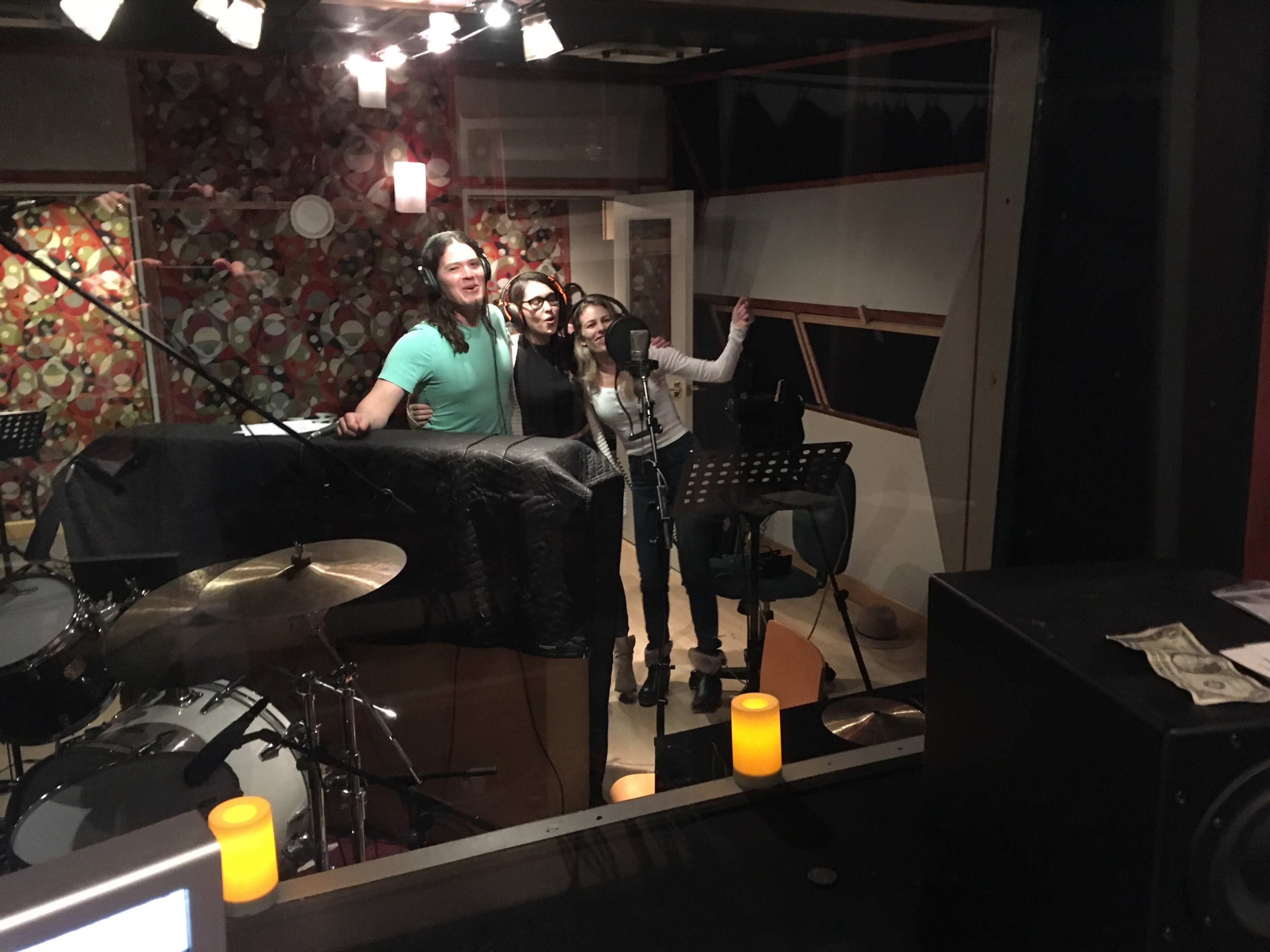 Three people stand behind a microphone in a recording studio while a drum set is visible in the foreground.
