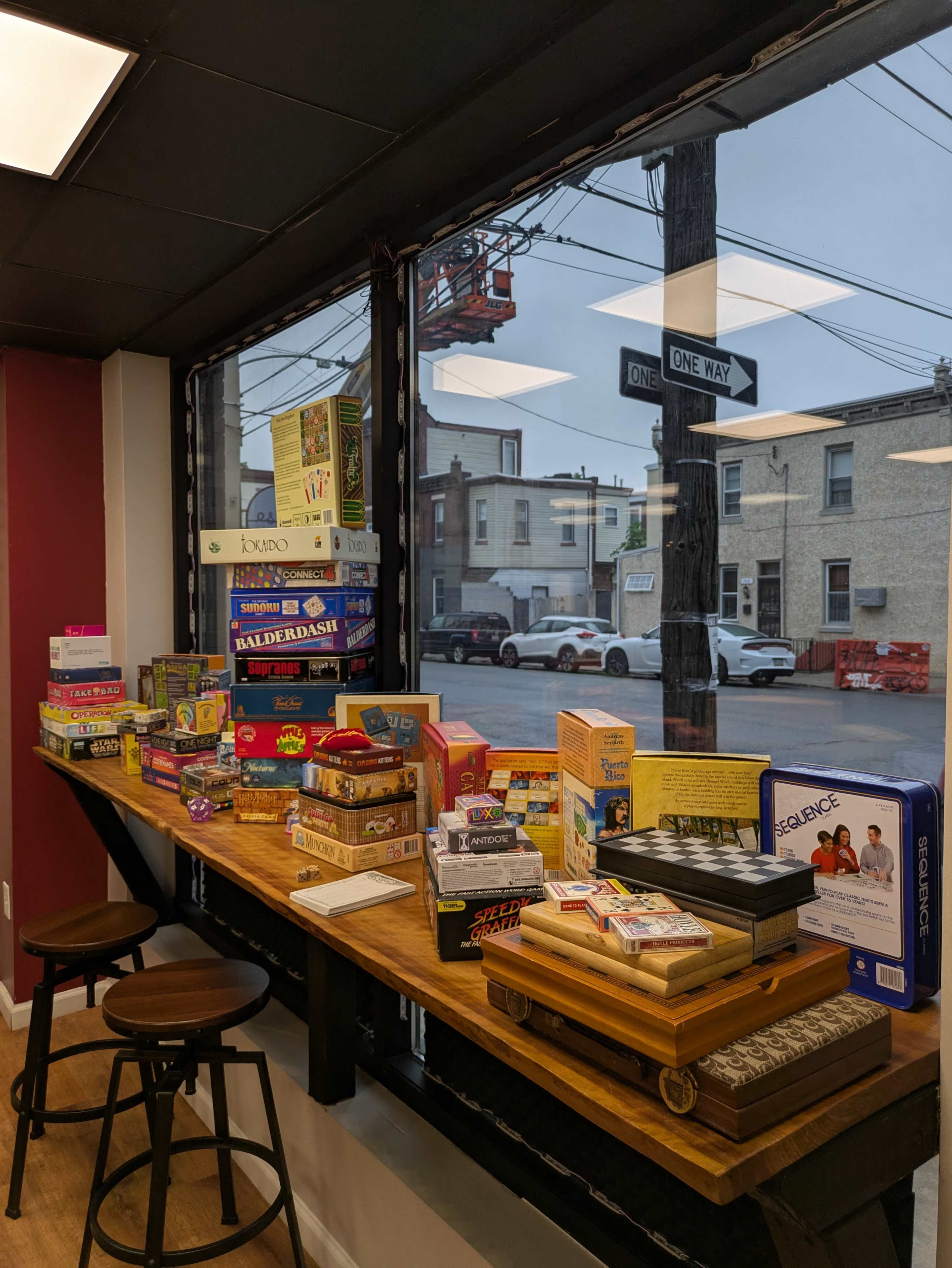 A wooden table displays various board games near a large window overlooking a street with parked cars and buildings.