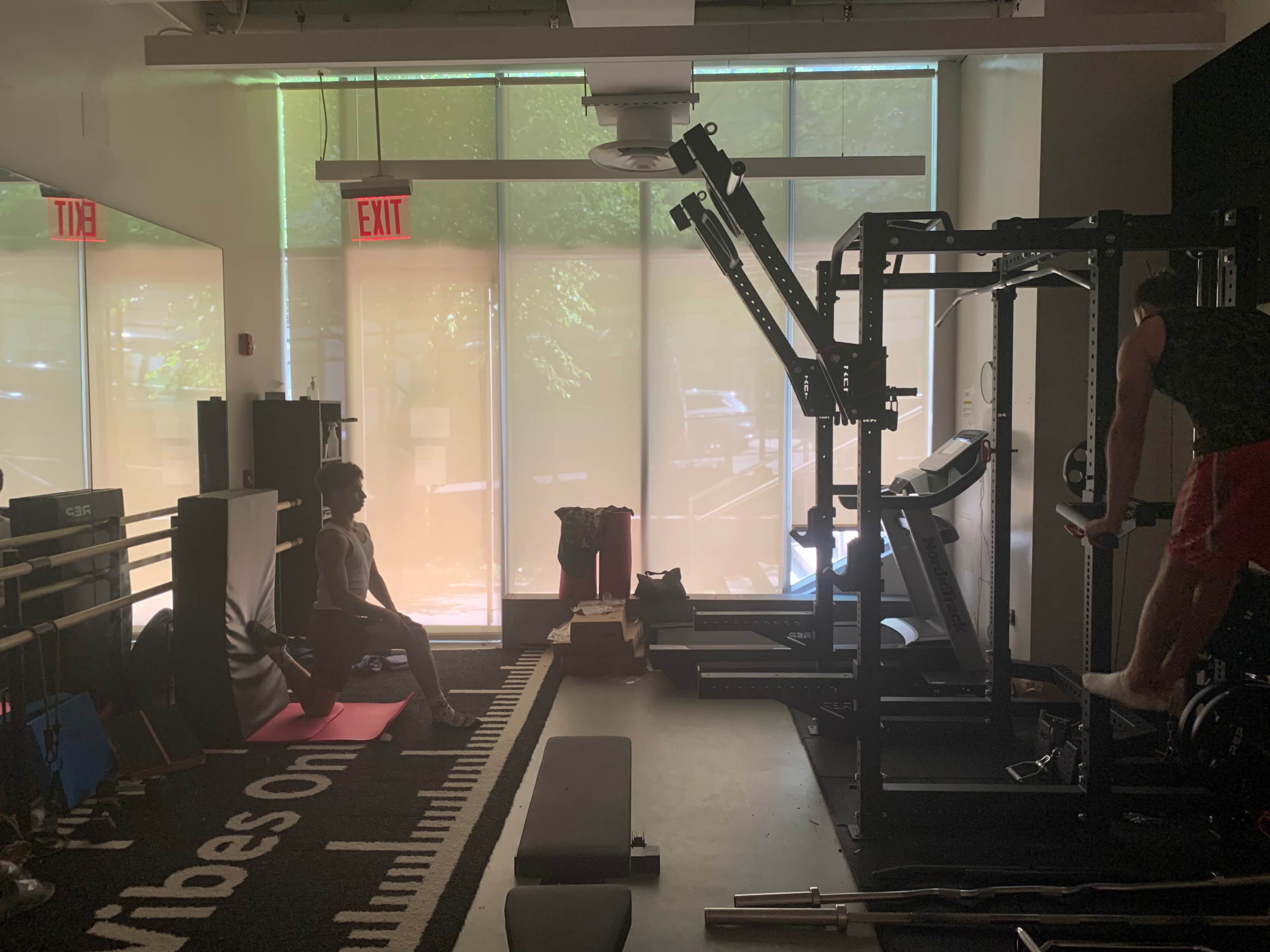 The image shows an indoor gym with exercise equipment, including a weightlifting rack, benches, and a person exercising while another sits nearby.
