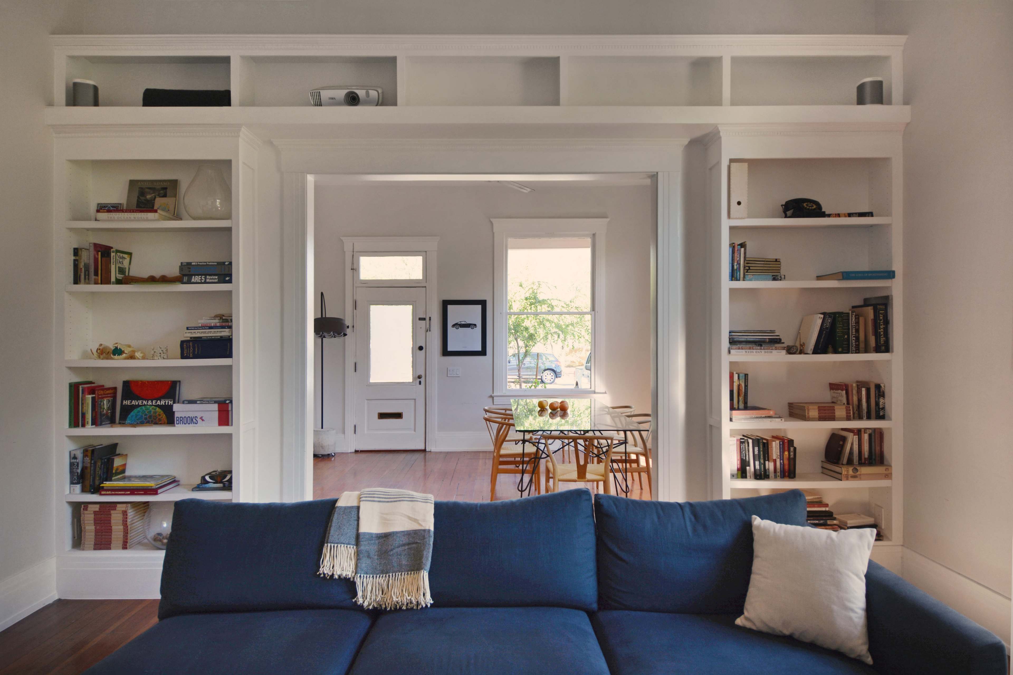 A living room features a large blue sofa facing built-in bookshelves, with a doorway leading to a dining area visible in the background.