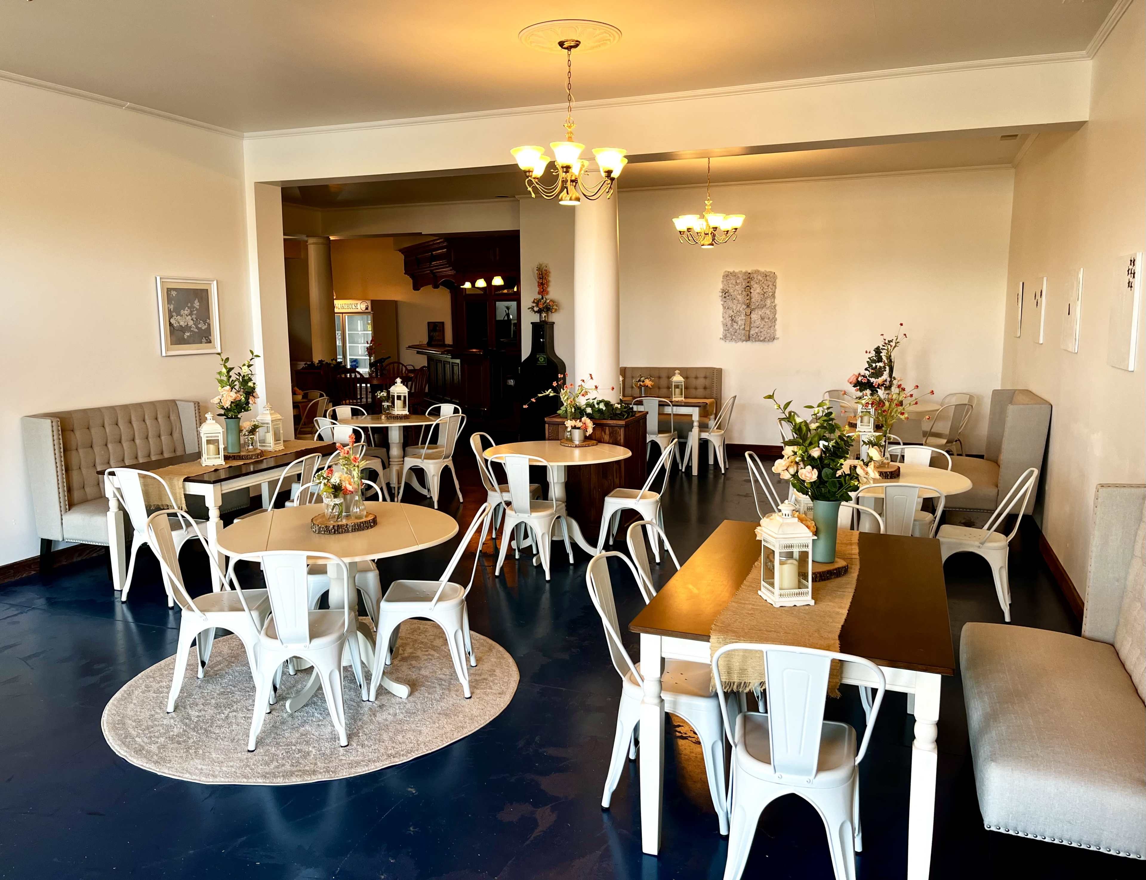 The image shows an indoor dining area with several tables set for guests, featuring floral centerpieces and a light-colored decor.