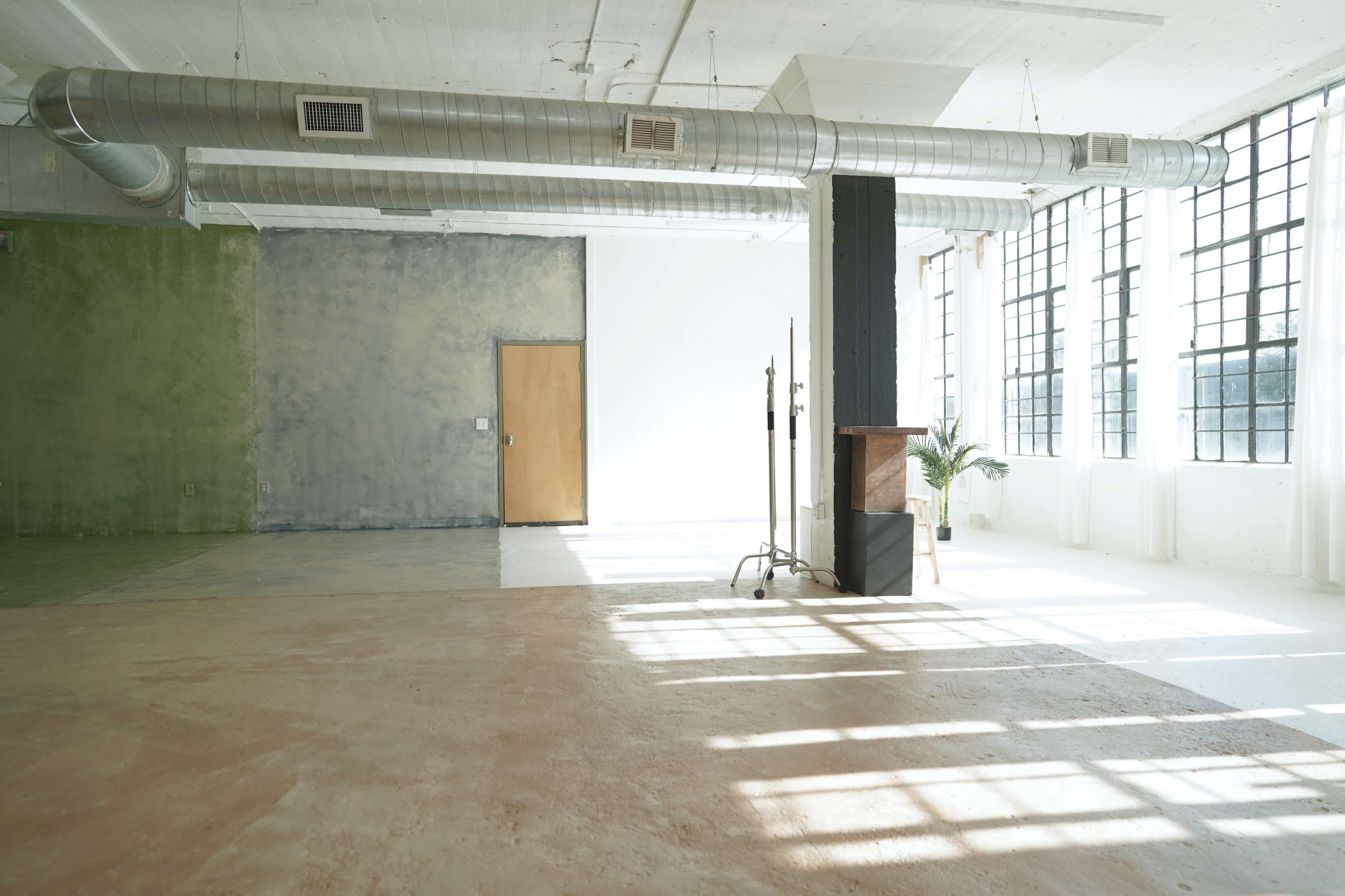 The image shows a spacious, industrial-style room with large windows, concrete floors, and a combination of green and gray textured walls.
