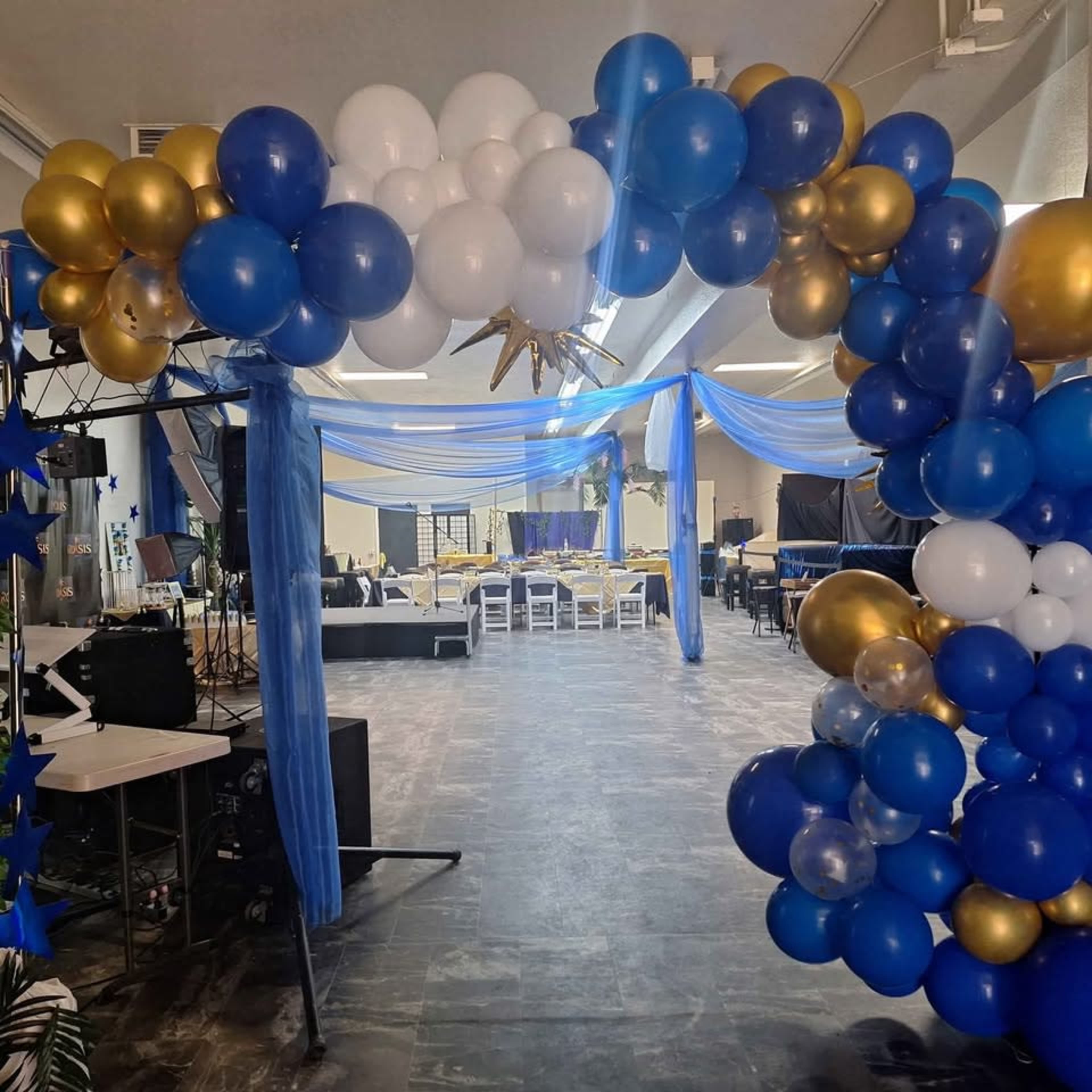 A balloon arch in blue, gold, and white colors welcomes guests into a decorated event space featuring tables and draped fabric.