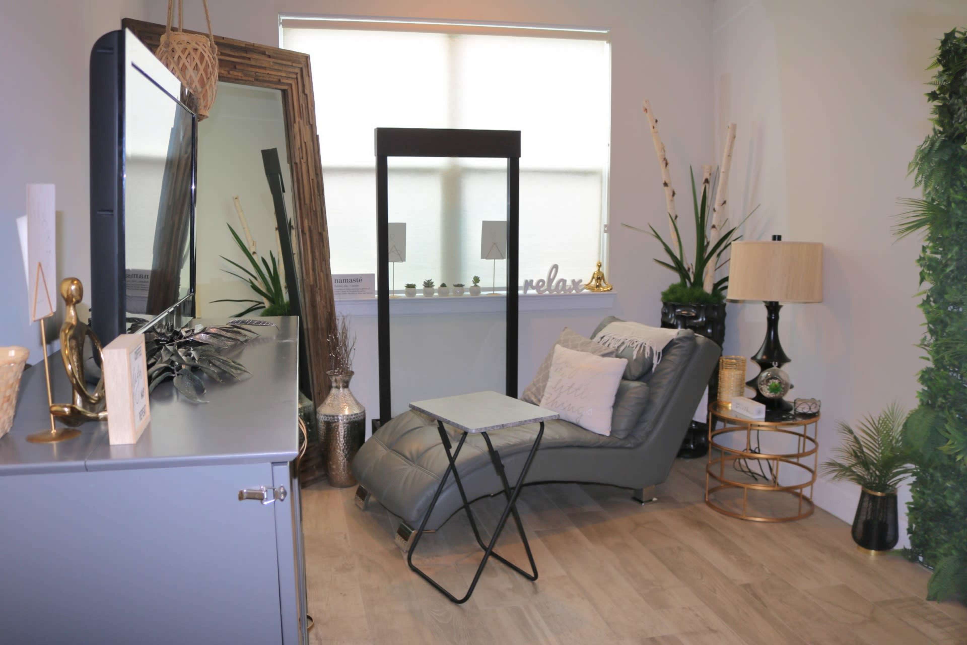 The image shows a cozy interior space with a gray reclining chair, a small side table, a decorative mirror, and various plants and decorative items arranged around the room.