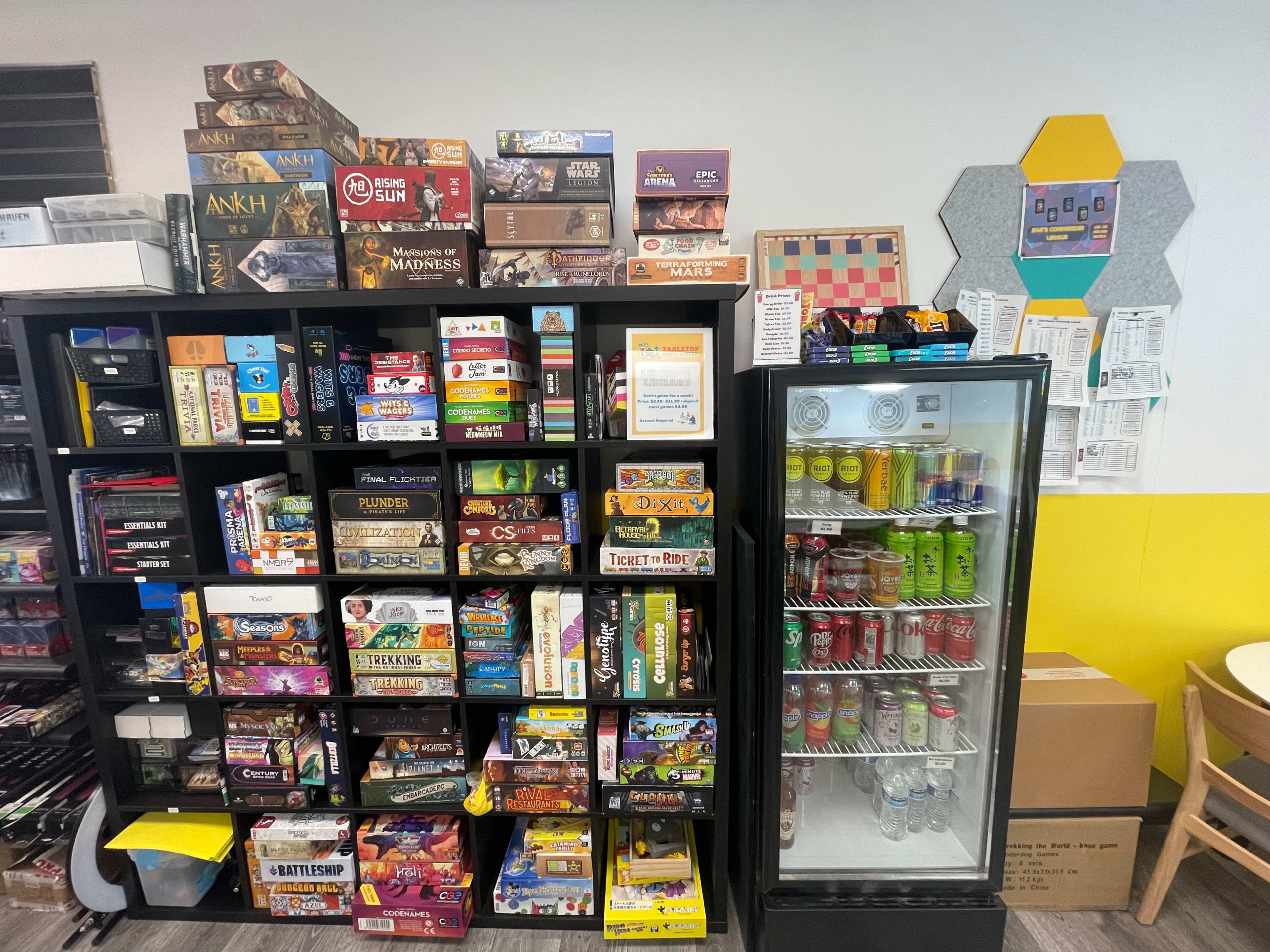 A shelving unit filled with various board games next to a glass-front refrigerator stocked with drinks.