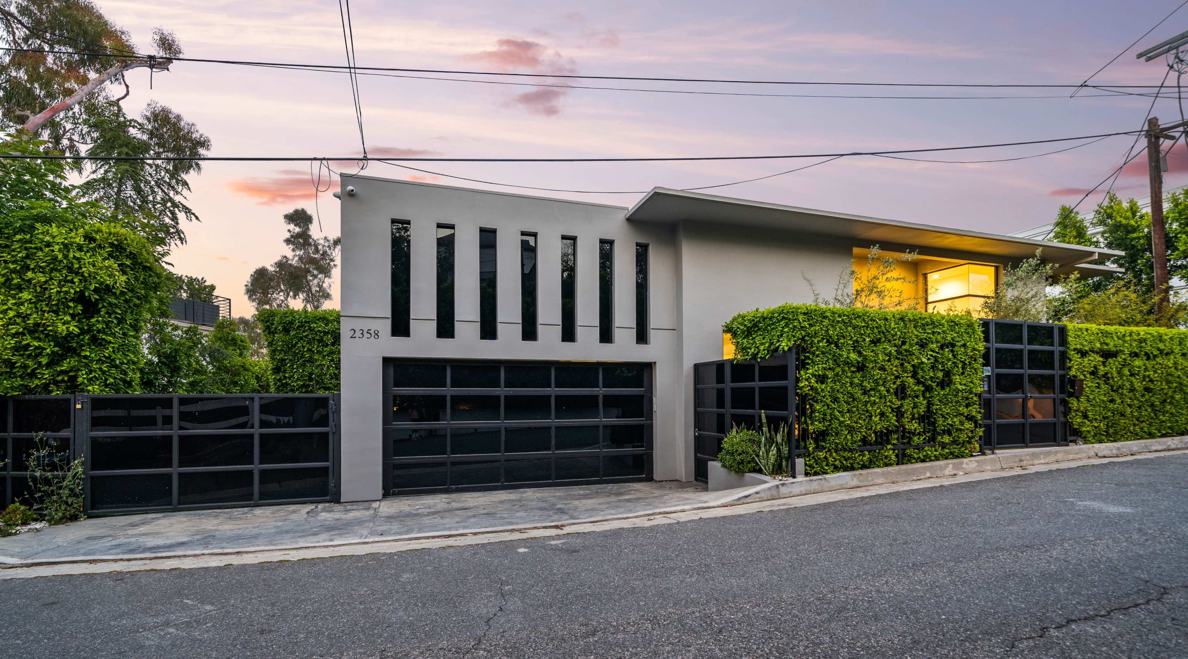 A modern, two-story house with large black windows and a garage door is flanked by neatly trimmed greenery on a quiet street.