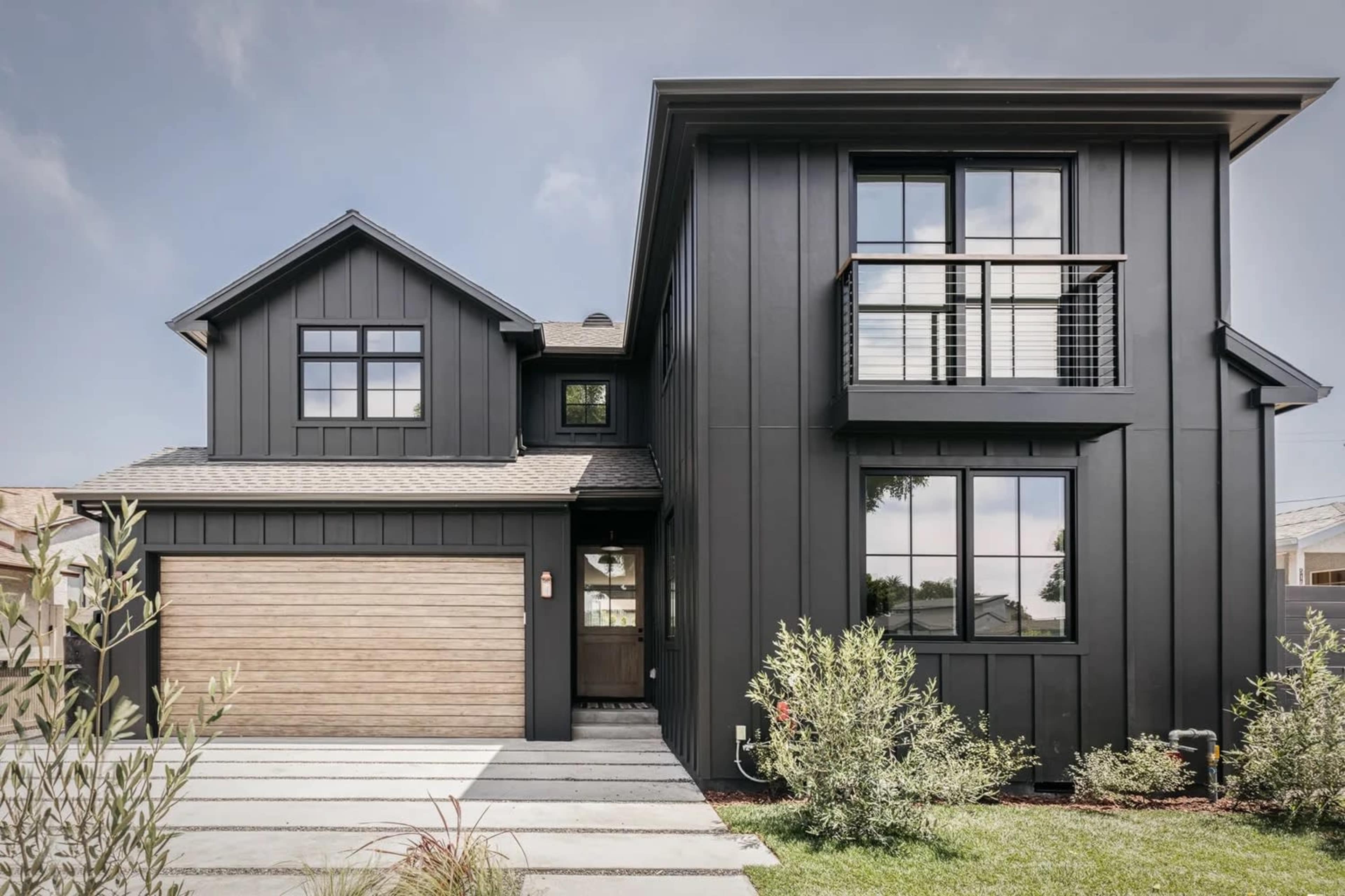 The image shows a modern two-story black house with a wooden garage door and large windows, framed by landscaped greenery.
