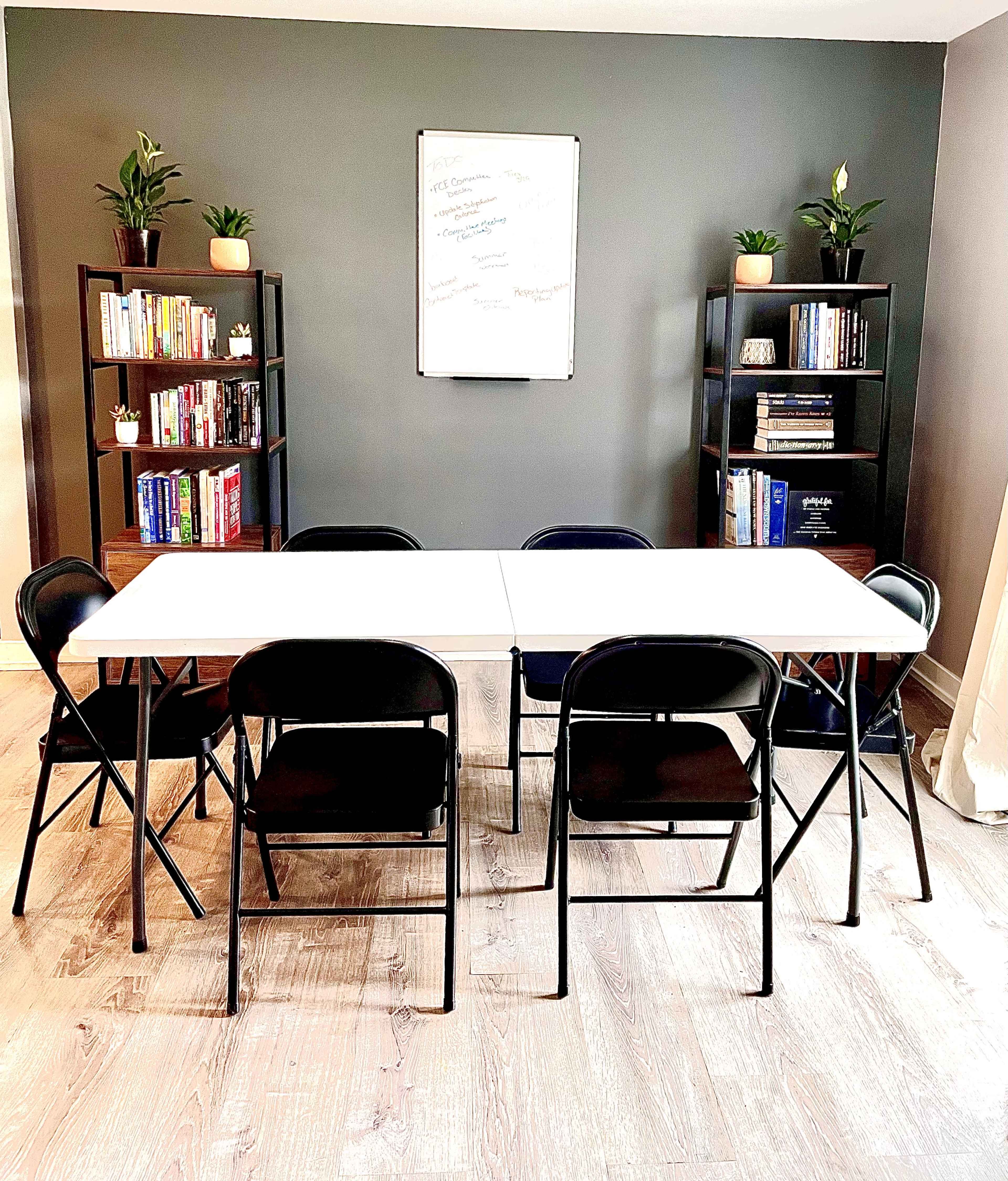 A rectangular table with six folding chairs arranged around it, set in a room featuring two bookshelves filled with books, potted plants, and a whiteboard on the wall.