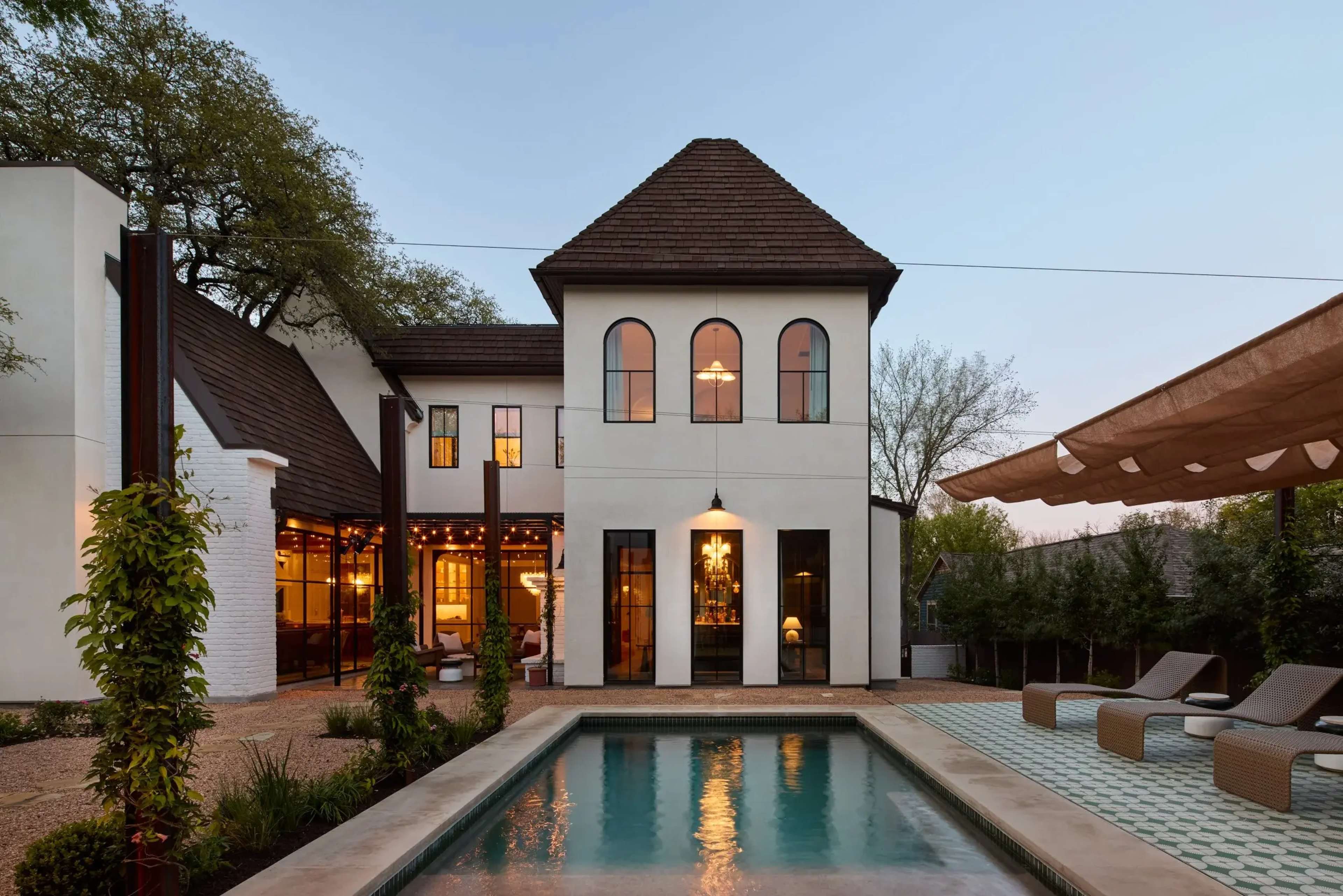 A modern house with a distinctive tower features a swimming pool and outdoor seating area surrounded by landscaped greenery.
