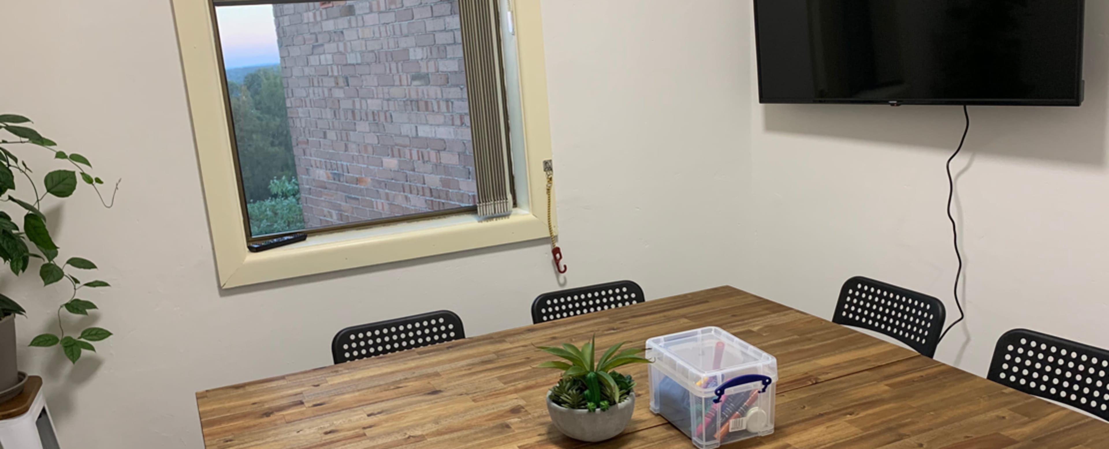 A meeting room features a wooden table with a potted plant and a storage container, along with a television mounted on the wall and a window showing a view outside.