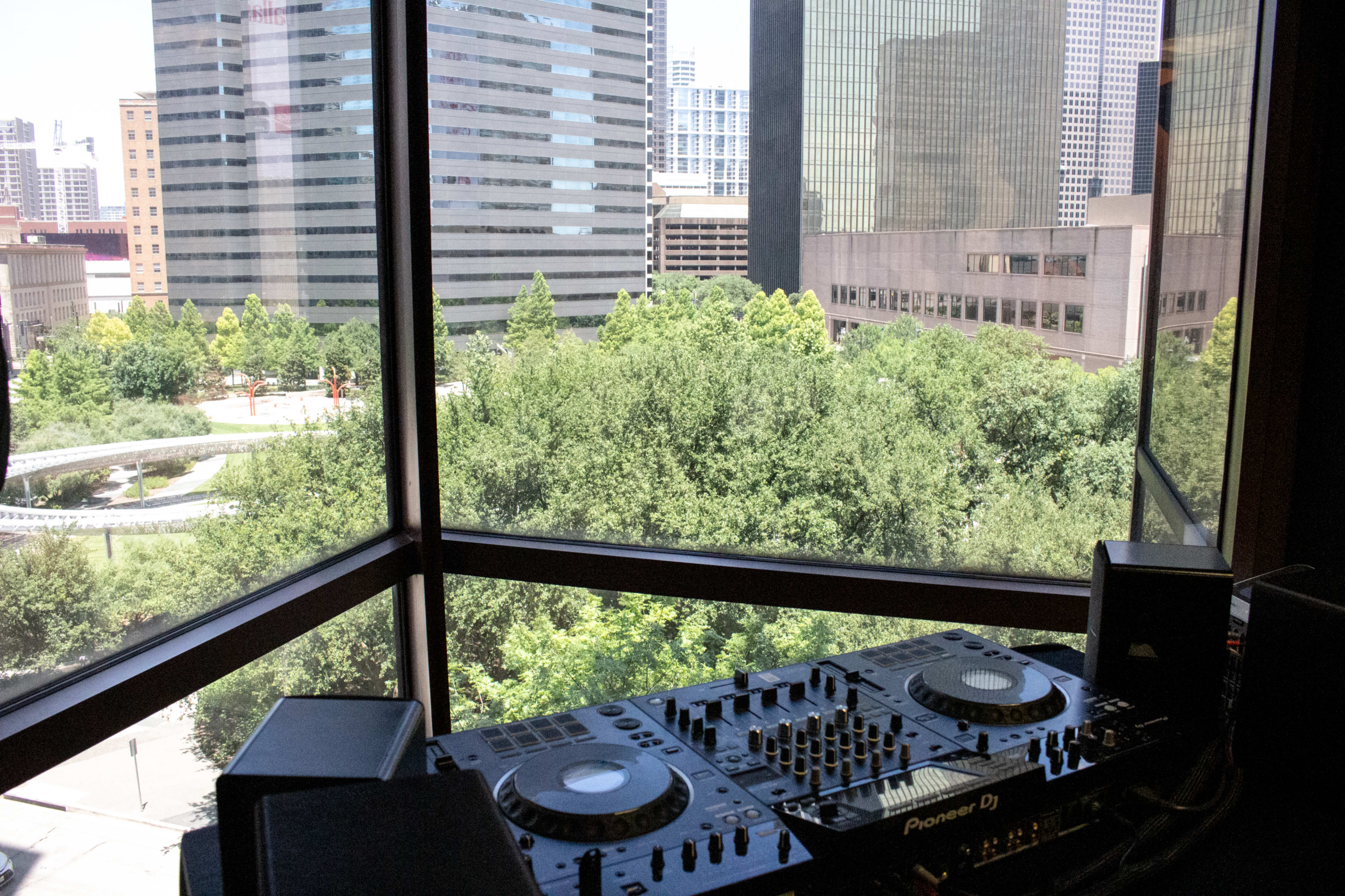 The image shows a DJ setup with two turntables positioned near a large window overlooking a green park and city skyline.