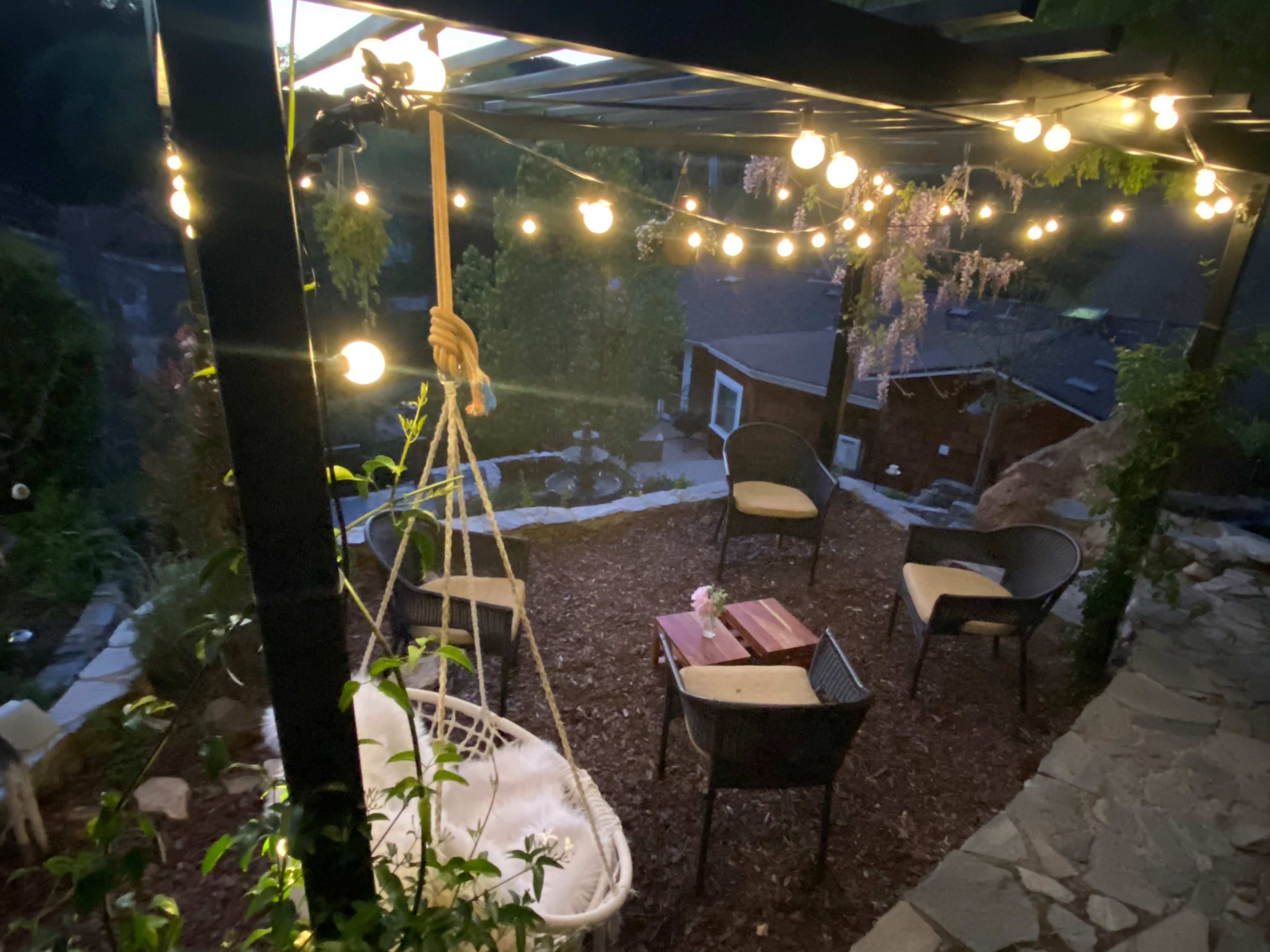 A cozy outdoor seating area features a hanging swing, string lights, and several chairs arranged around a wooden table.
