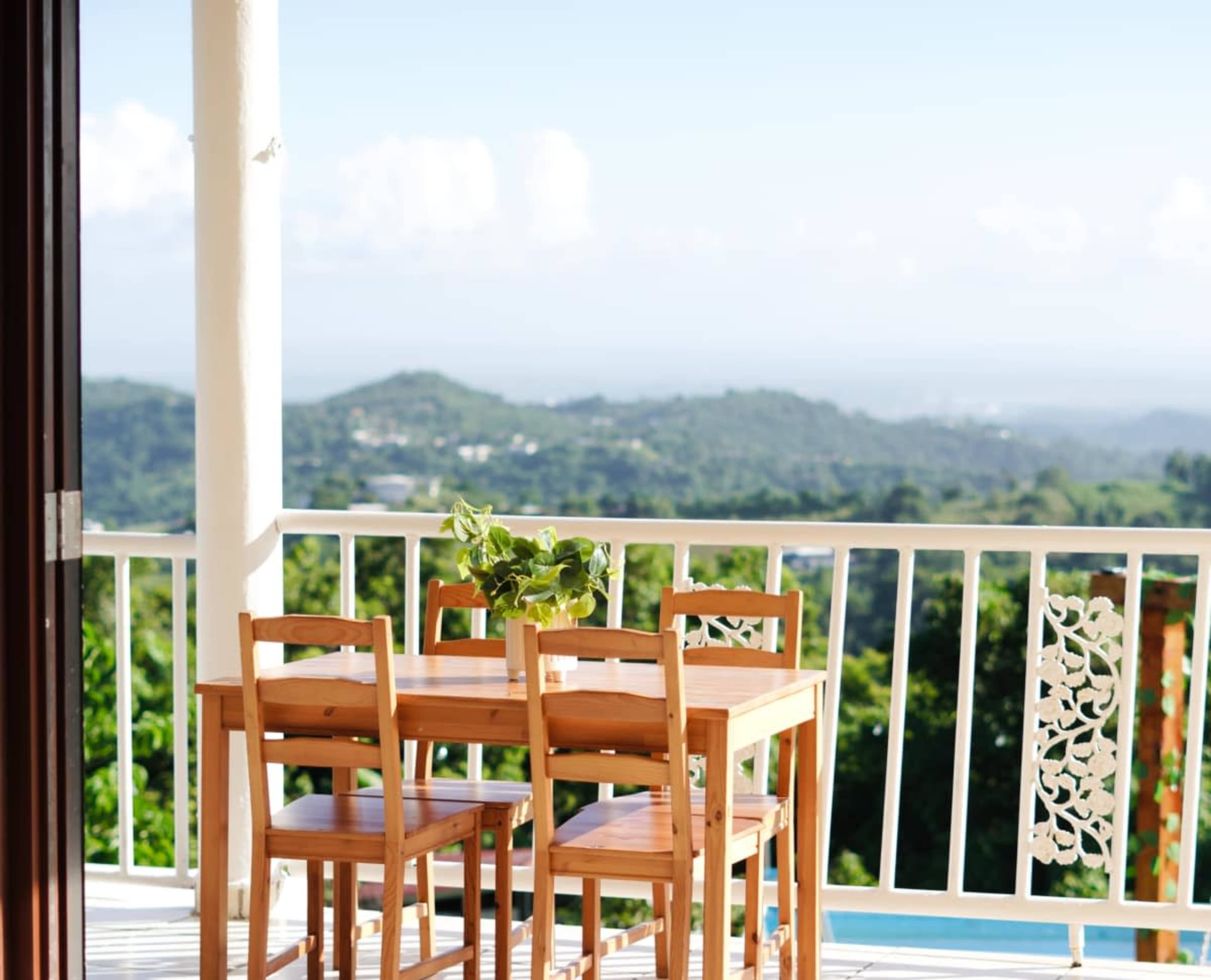 A wooden dining table and chairs sit on a balcony overlooking a green hillside under a clear blue sky.