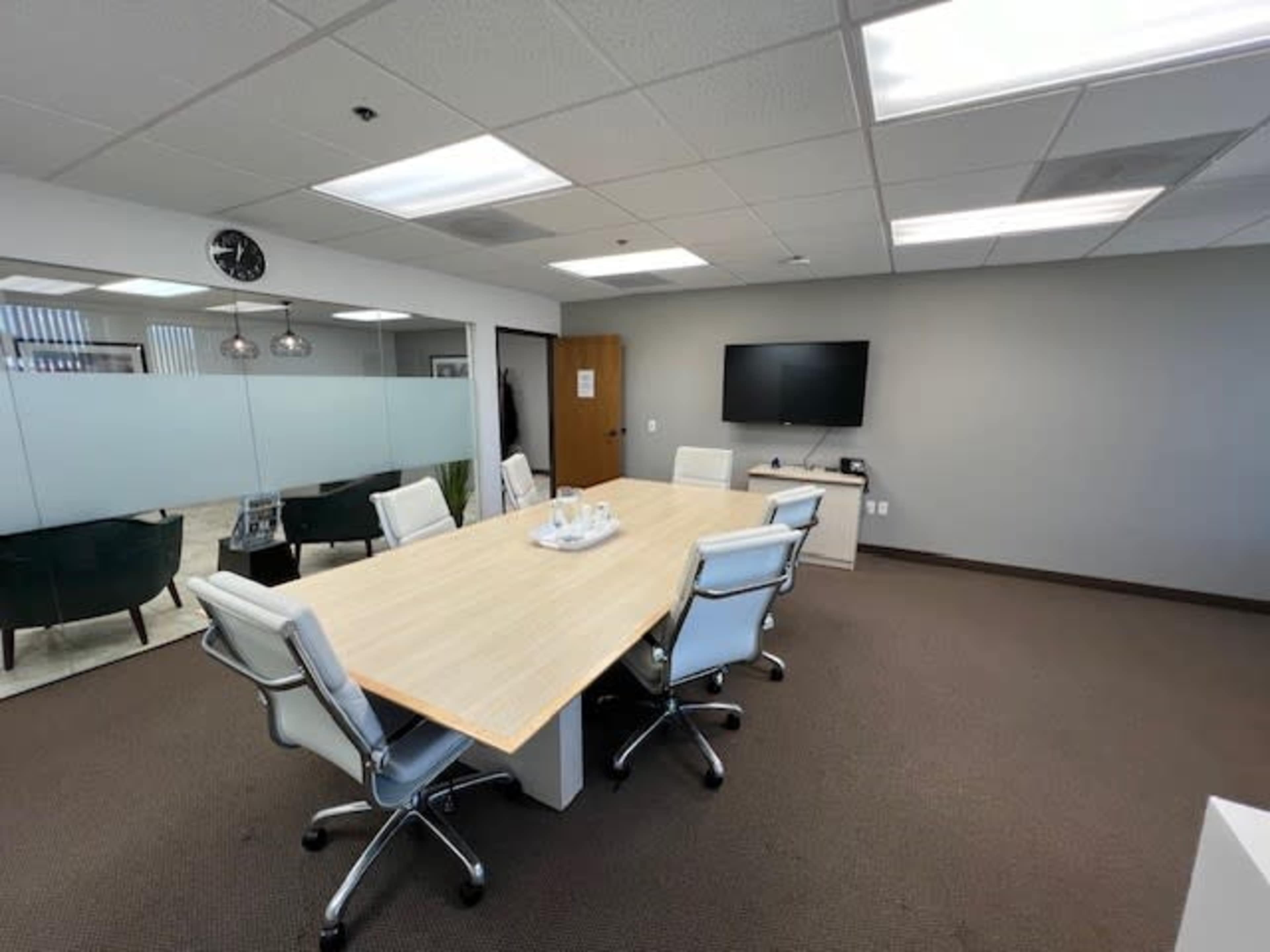 A conference room features a large wooden table surrounded by six white office chairs, with a television mounted on the wall and glass partitions.