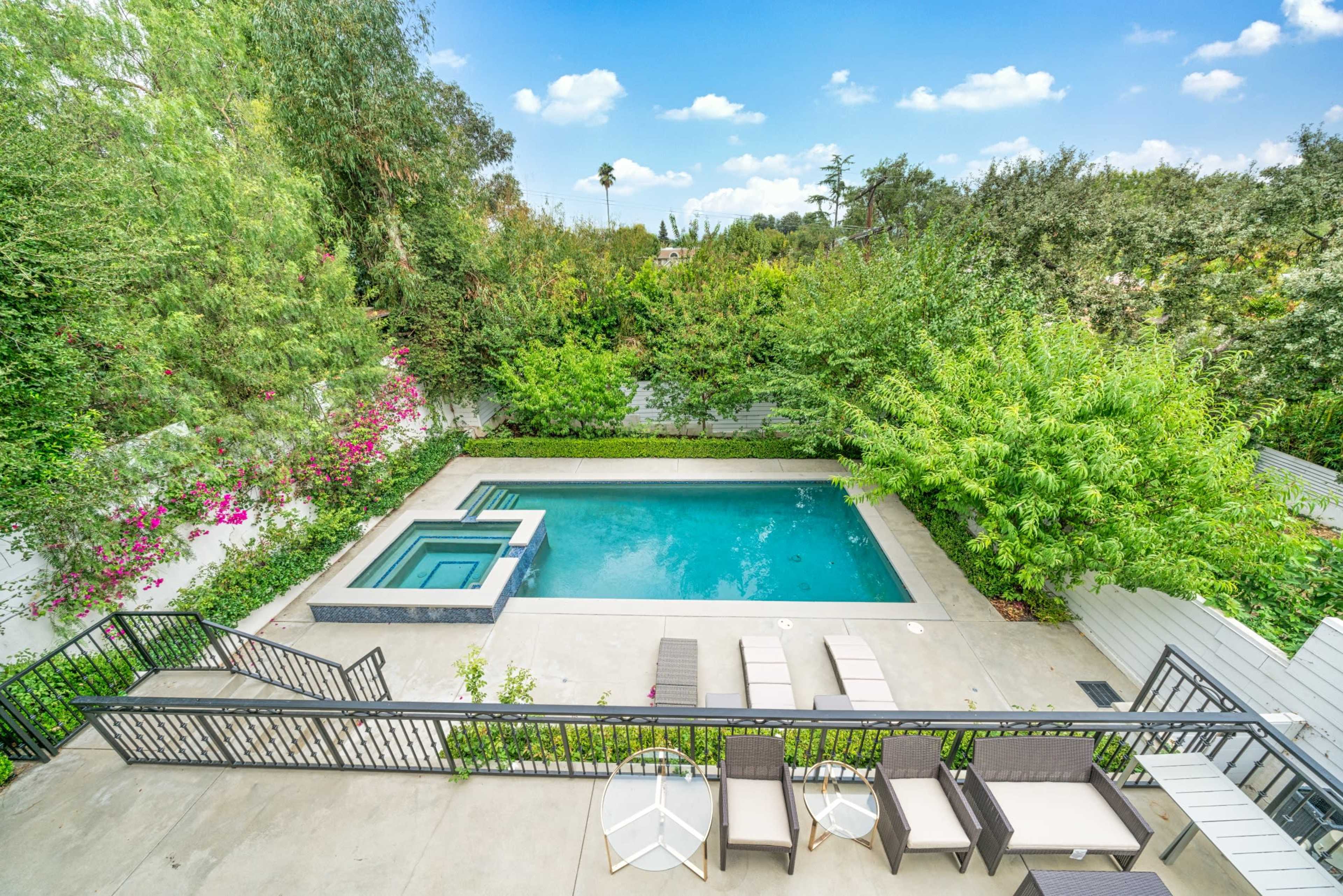 The image shows a backyard with a swimming pool and spa surrounded by lush greenery and a patio area with lounge chairs.