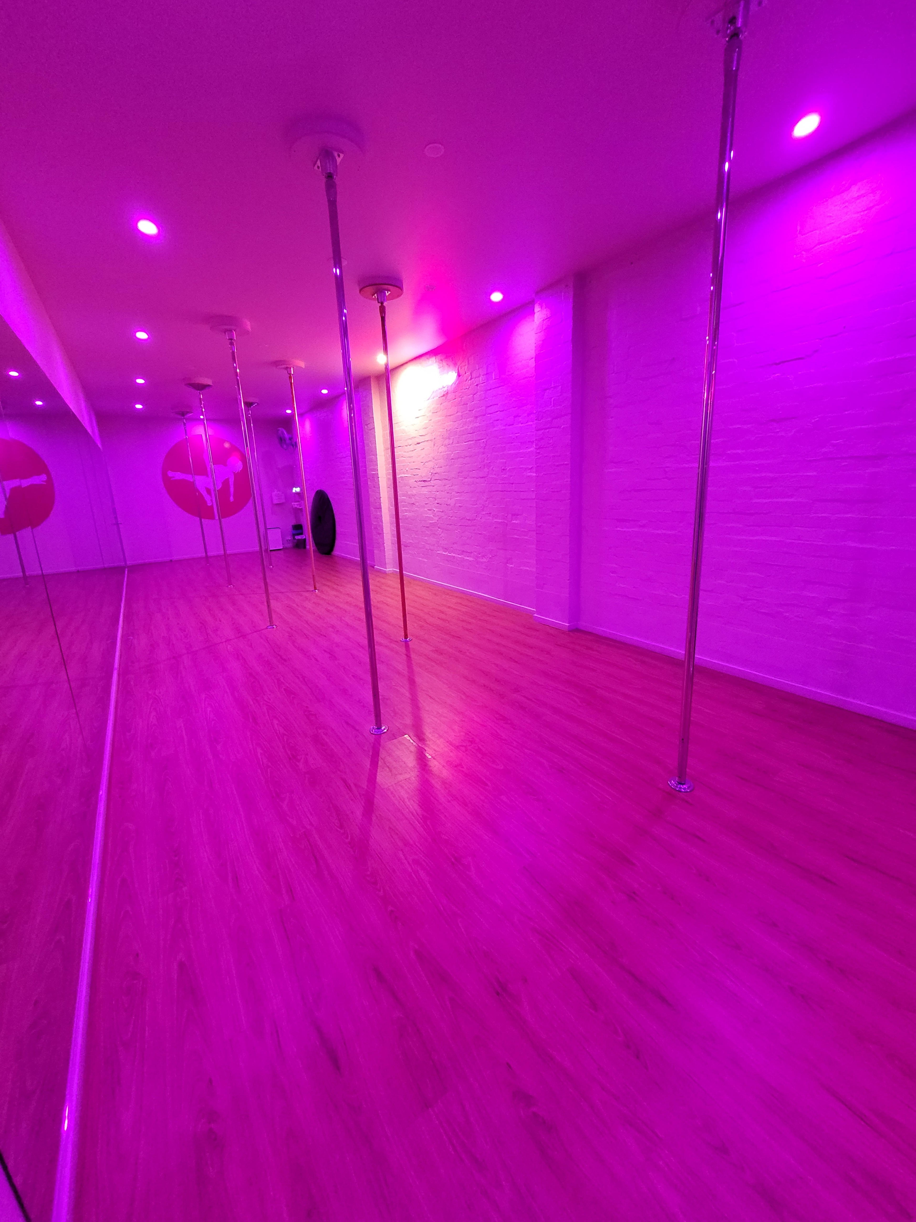 A dance studio with wooden flooring, several poles, and purple ambient lighting.