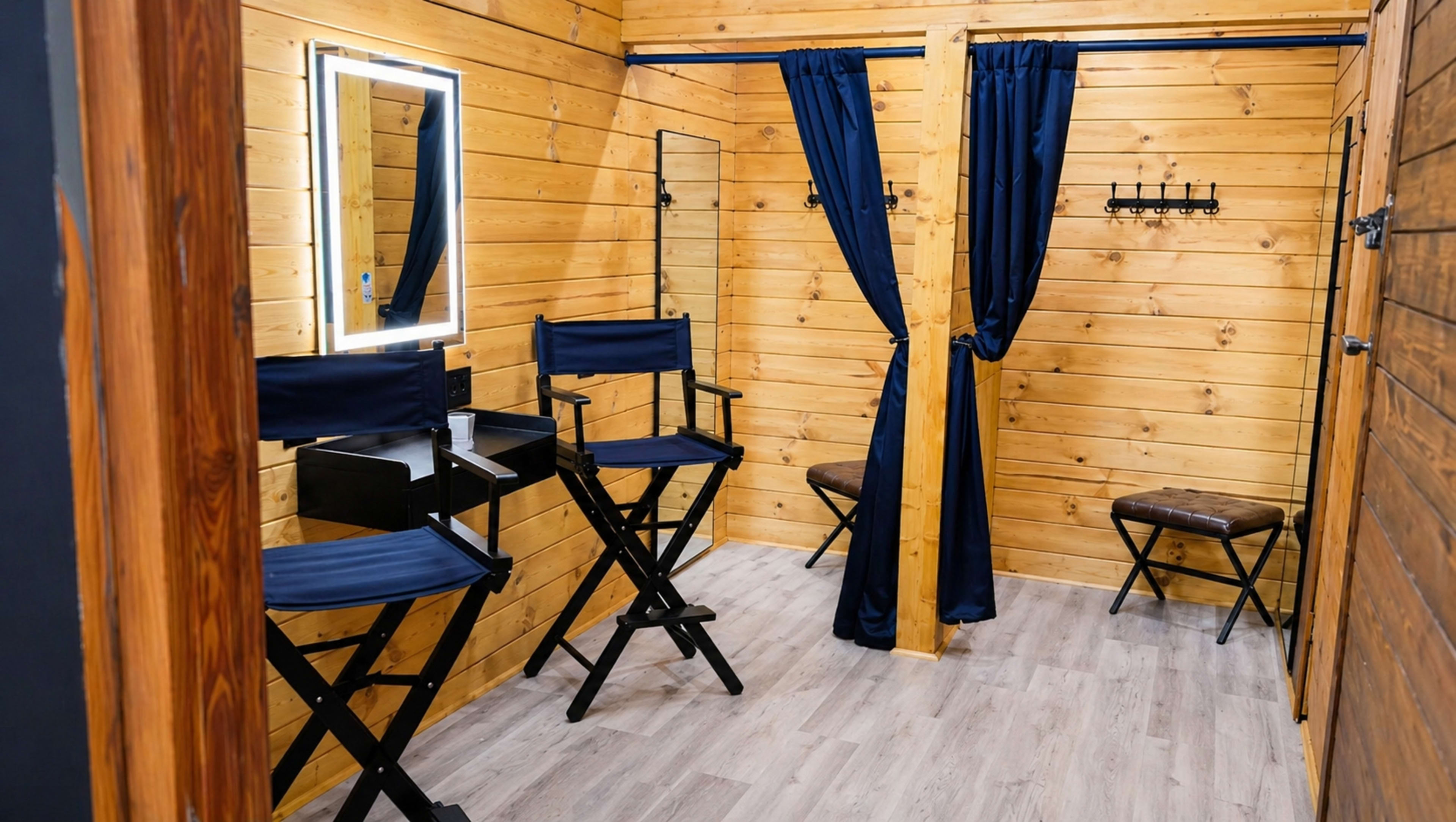The image shows a small room with wooden walls, featuring two tall director's chairs, a mirror with LED lighting, curtains, and a few additional seating options.