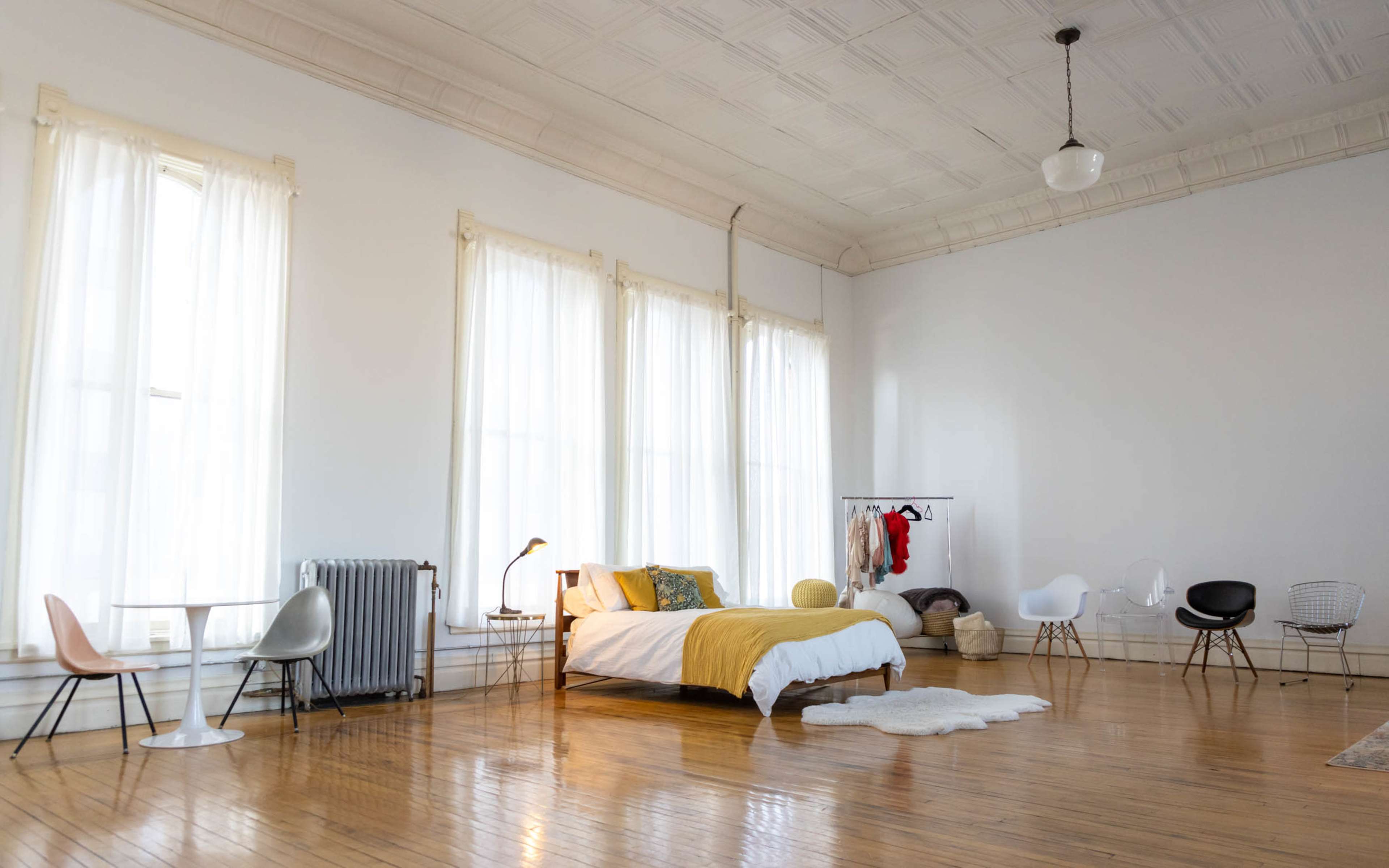 A spacious bedroom features a bed with yellow bedding, large windows draped in sheer curtains, and minimalist furniture including a table and various chairs.