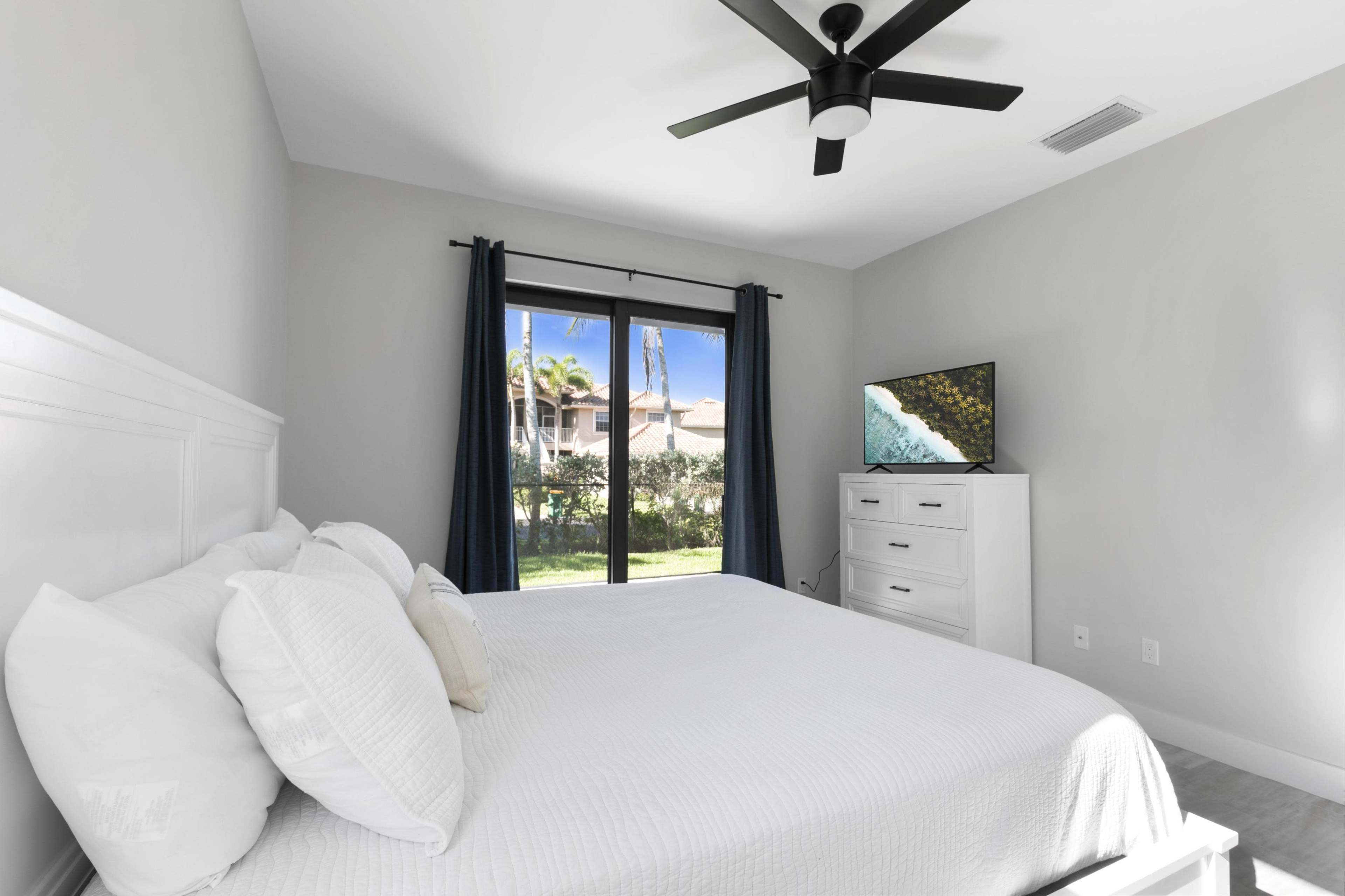 A bedroom features a white bed with decorative pillows, a dresser, a television, and large windows with green curtains overlooking a garden.
