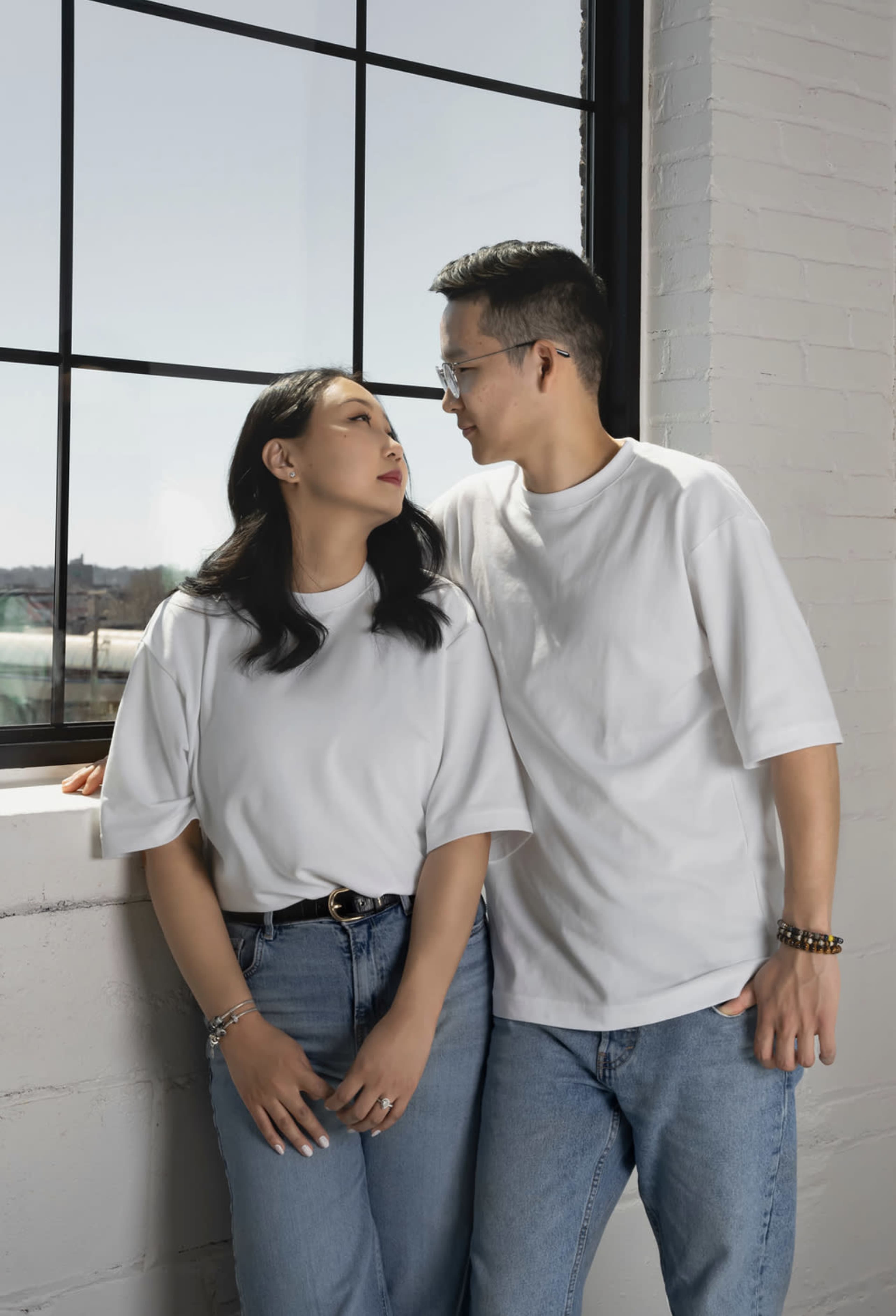 A couple wearing matching white t-shirts and denim jeans stands close together by a large window, gazing at each other.
