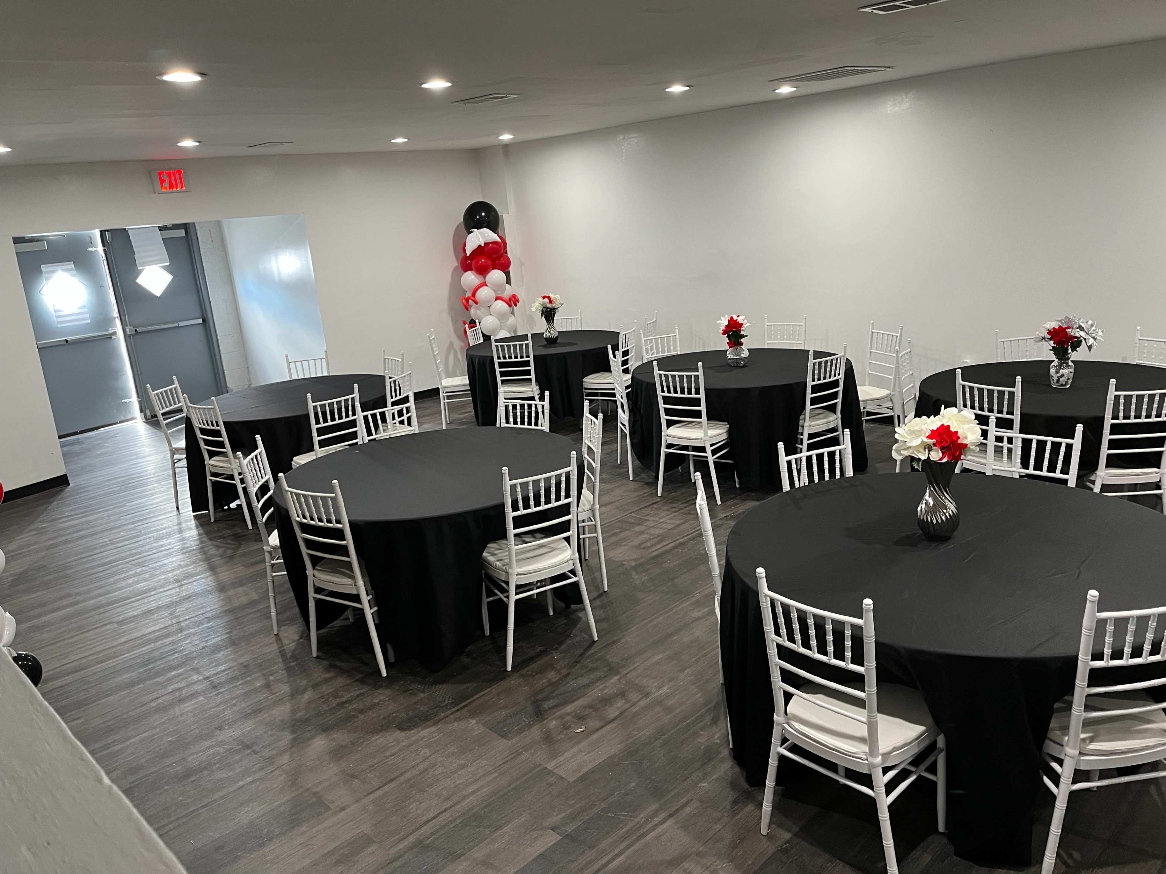 The image shows a spacious event room with several round tables covered in black tablecloths, each adorned with floral centerpieces, and a decorative balloon arch in the corner.