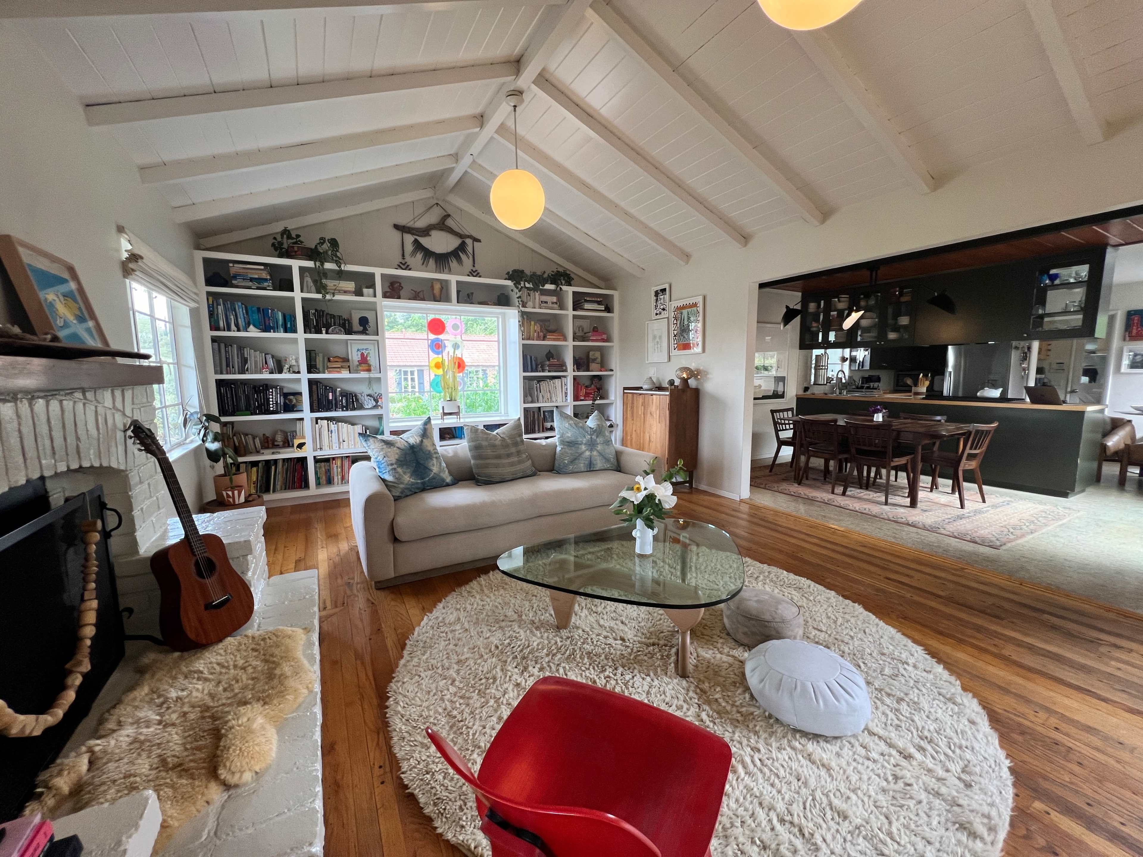 The scene shows a bright living room with a high, sloped ceiling, a glass coffee table on a circular rug, a bookshelf filled with books, and an open dining area in the background.