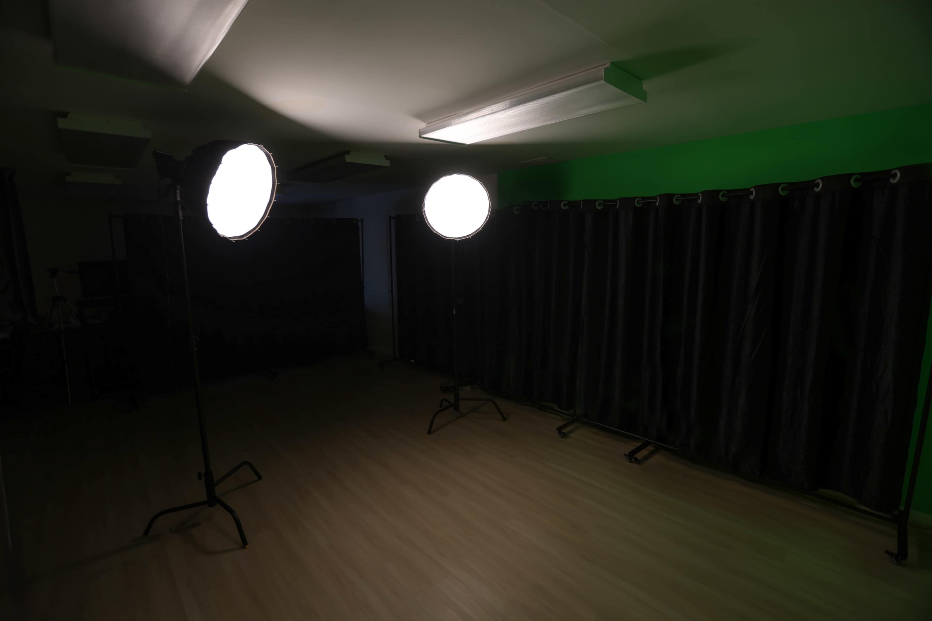 The image shows an empty studio space with two large lights on stands illuminating the area, alongside a black curtain and a green wall.