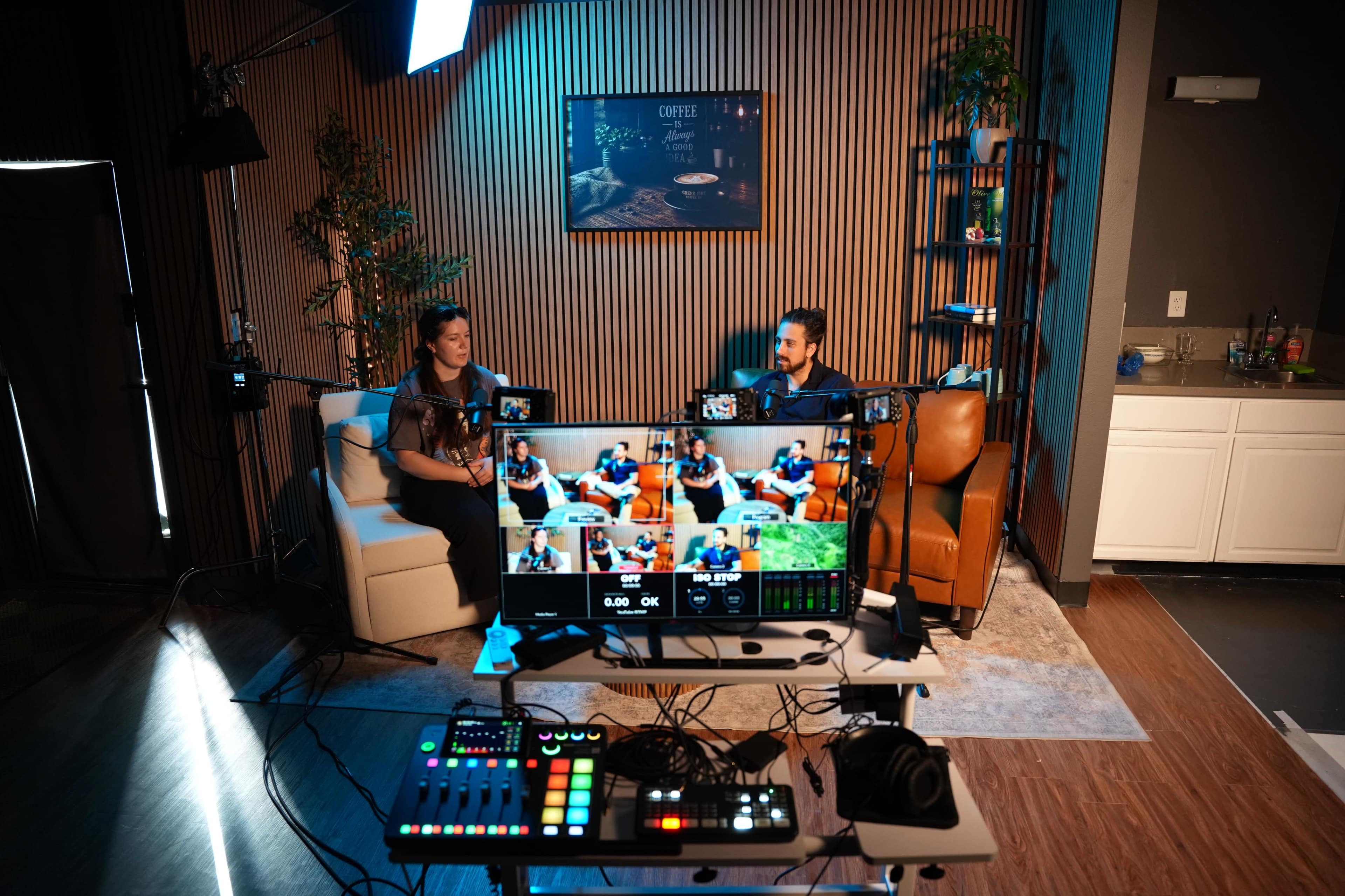 The image shows two men seated on a couch in a studio setup, surrounded by cameras and equipment, with a large screen displaying a live video feed in front of them.