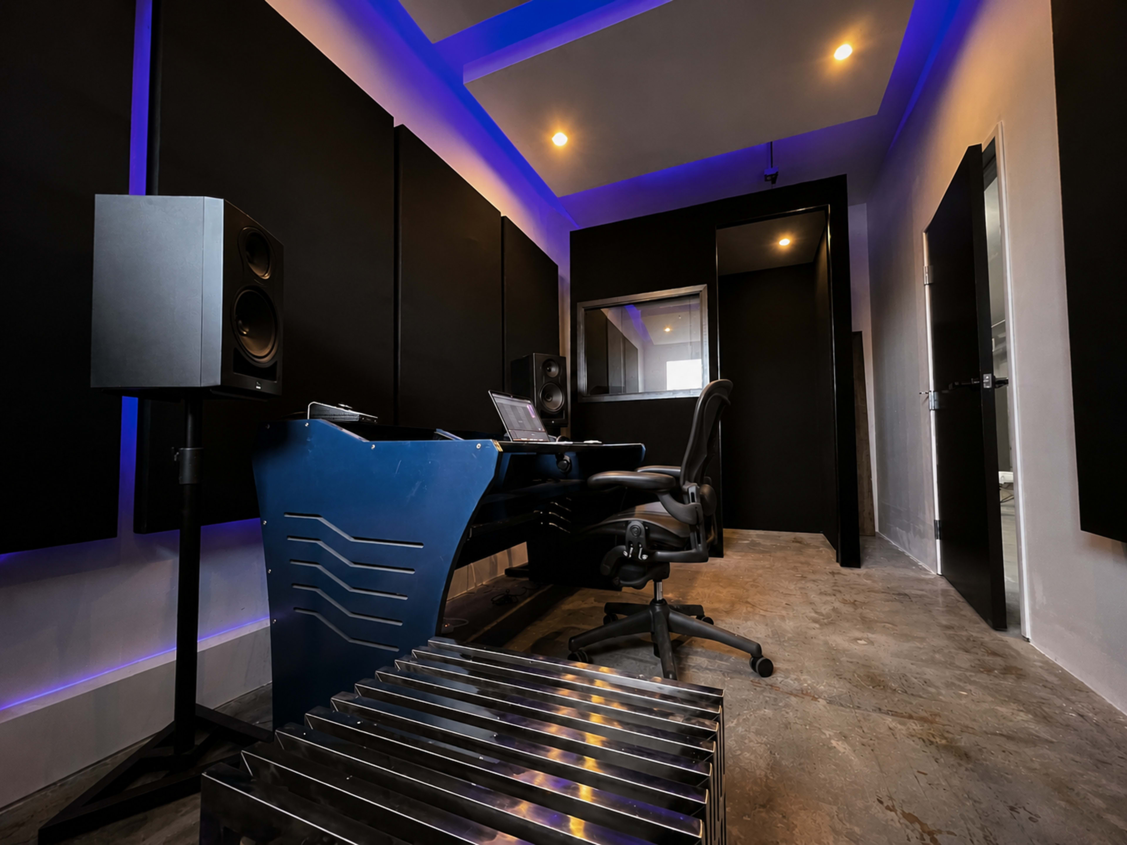The image shows a modern music studio with soundproof walls, a desk with a computer, and professional audio speakers.
