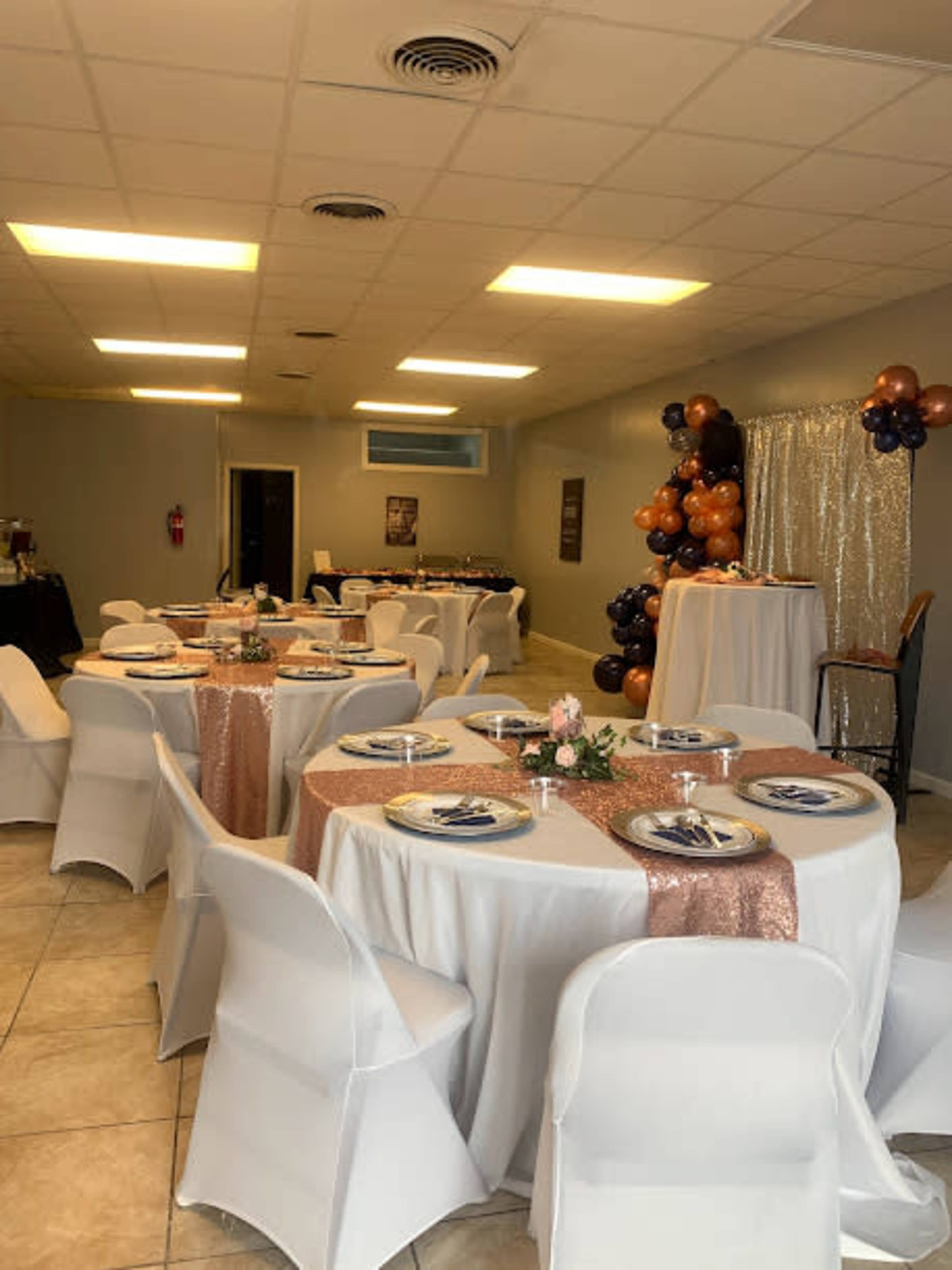 A banquet hall is set up with several tables covered in white tablecloths and decorated with rose gold accents, surrounded by chairs and a backdrop featuring balloons.