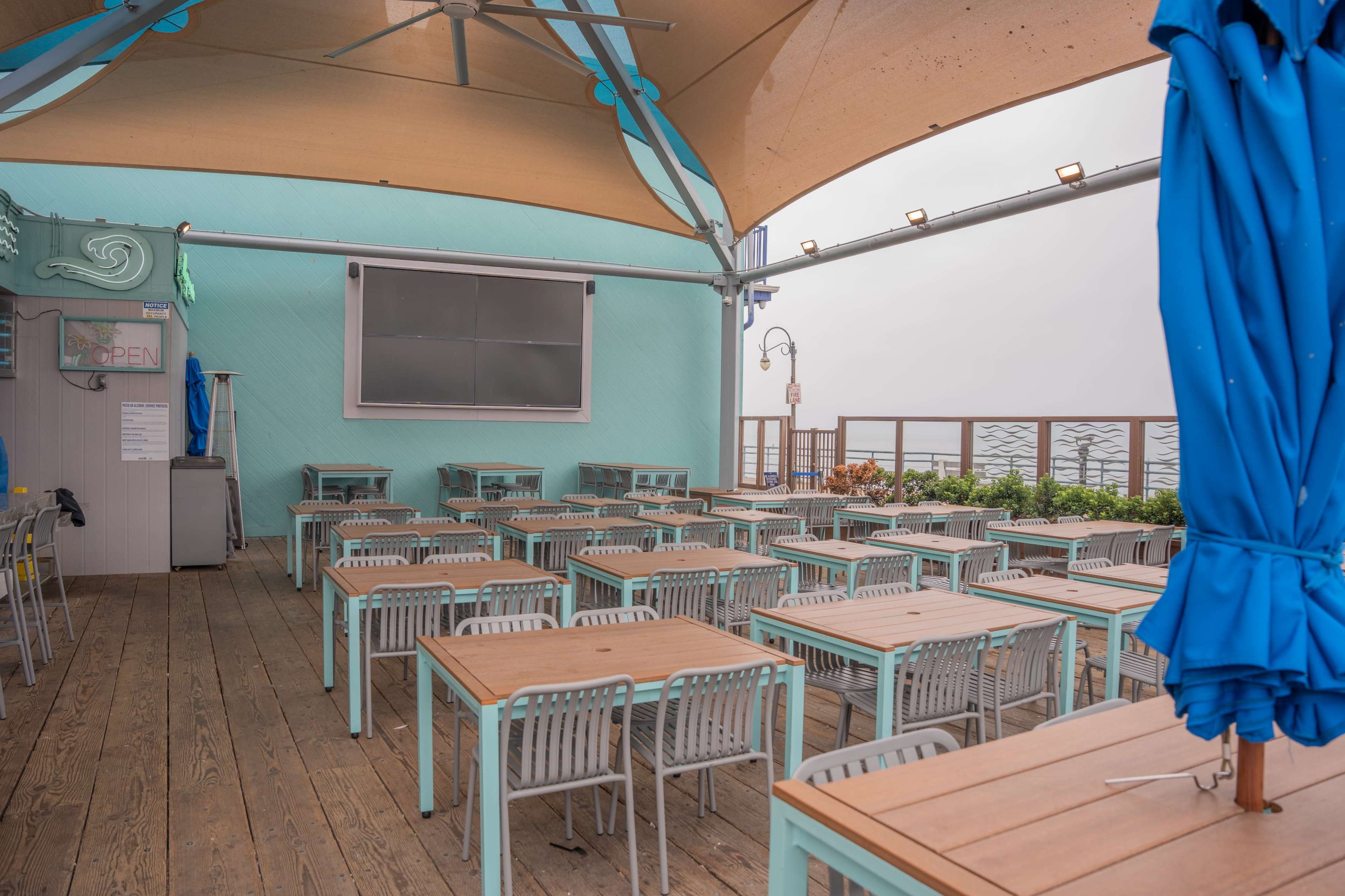 An outdoor seating area with wooden tables and chairs is covered by a large canopy in a coastal setting.