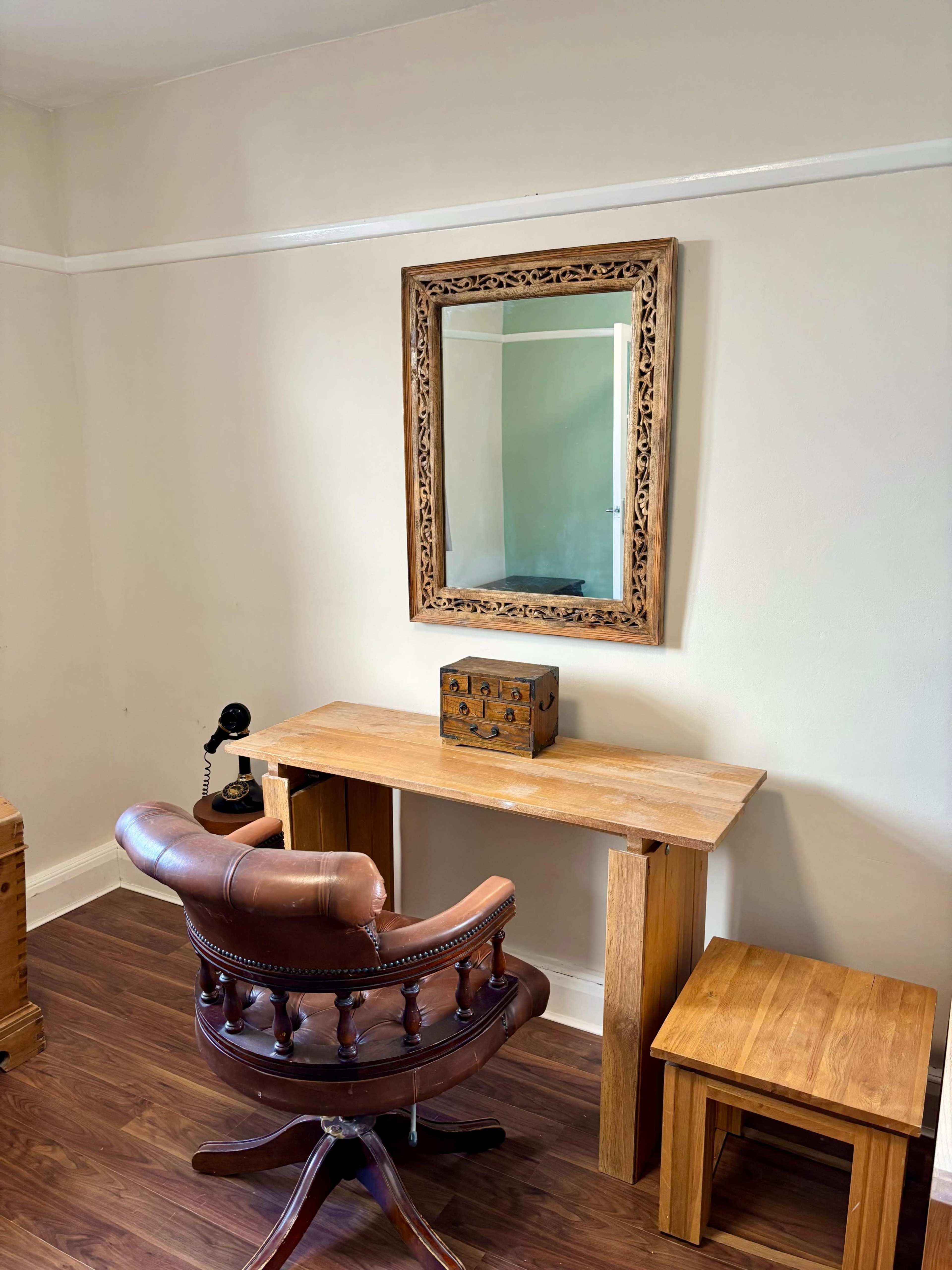 The image shows a wooden desk with a mirror above it, accompanied by a brown swivel chair and a small wooden stool in a minimalist room.
