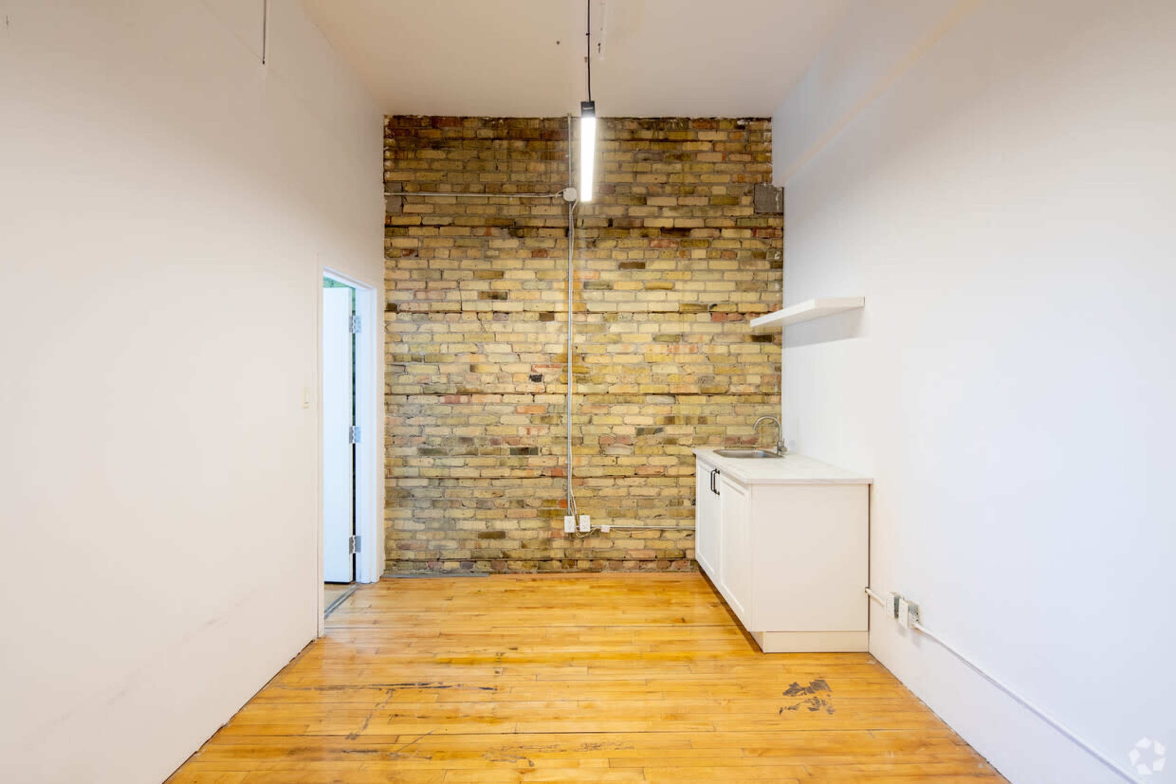 The image shows a small, empty room with a wooden floor, a brick wall, and a simple kitchenette along one side.