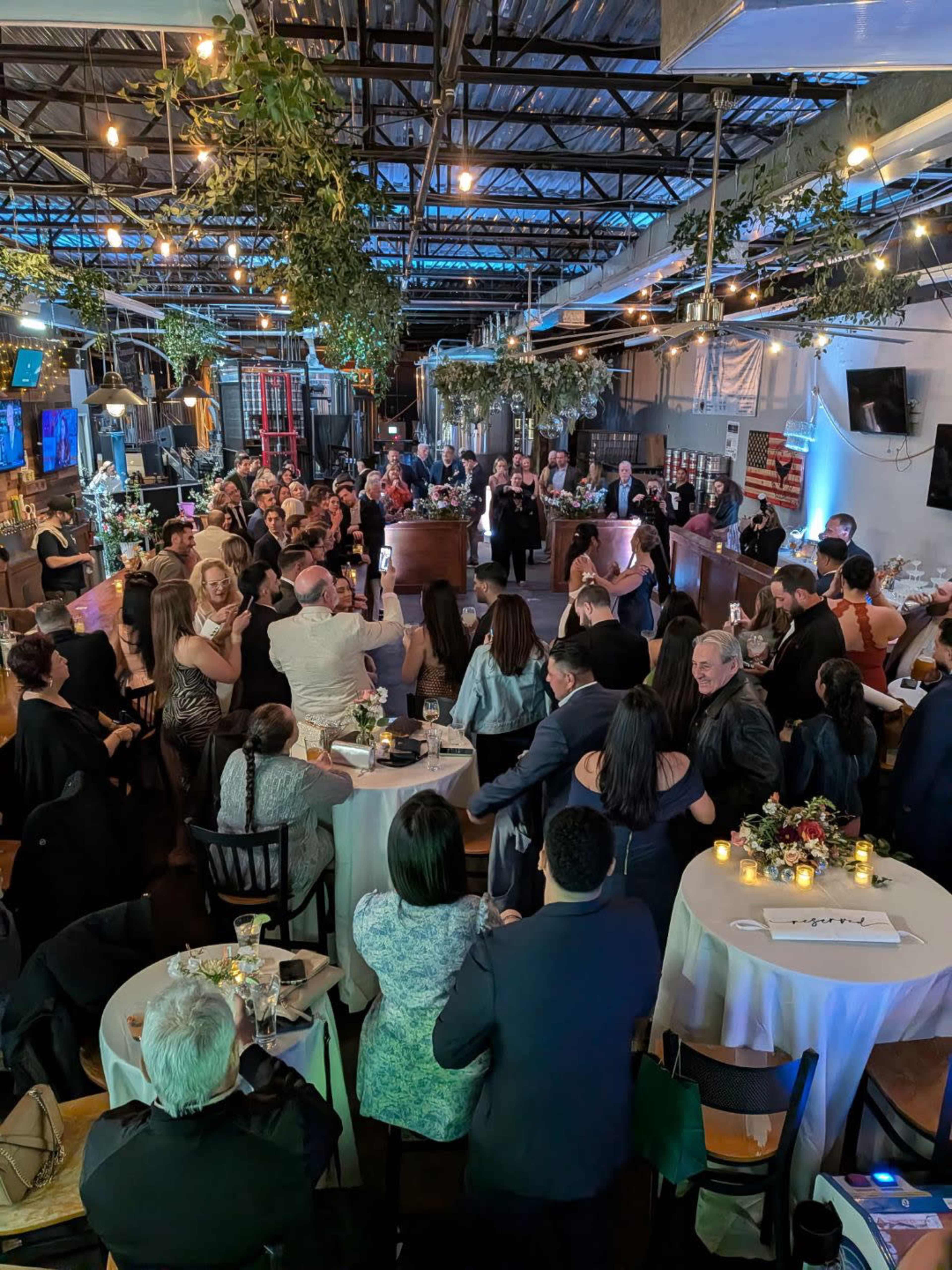 A large gathering of people is seated and standing around tables in an indoor event space decorated with greenery and soft lighting.