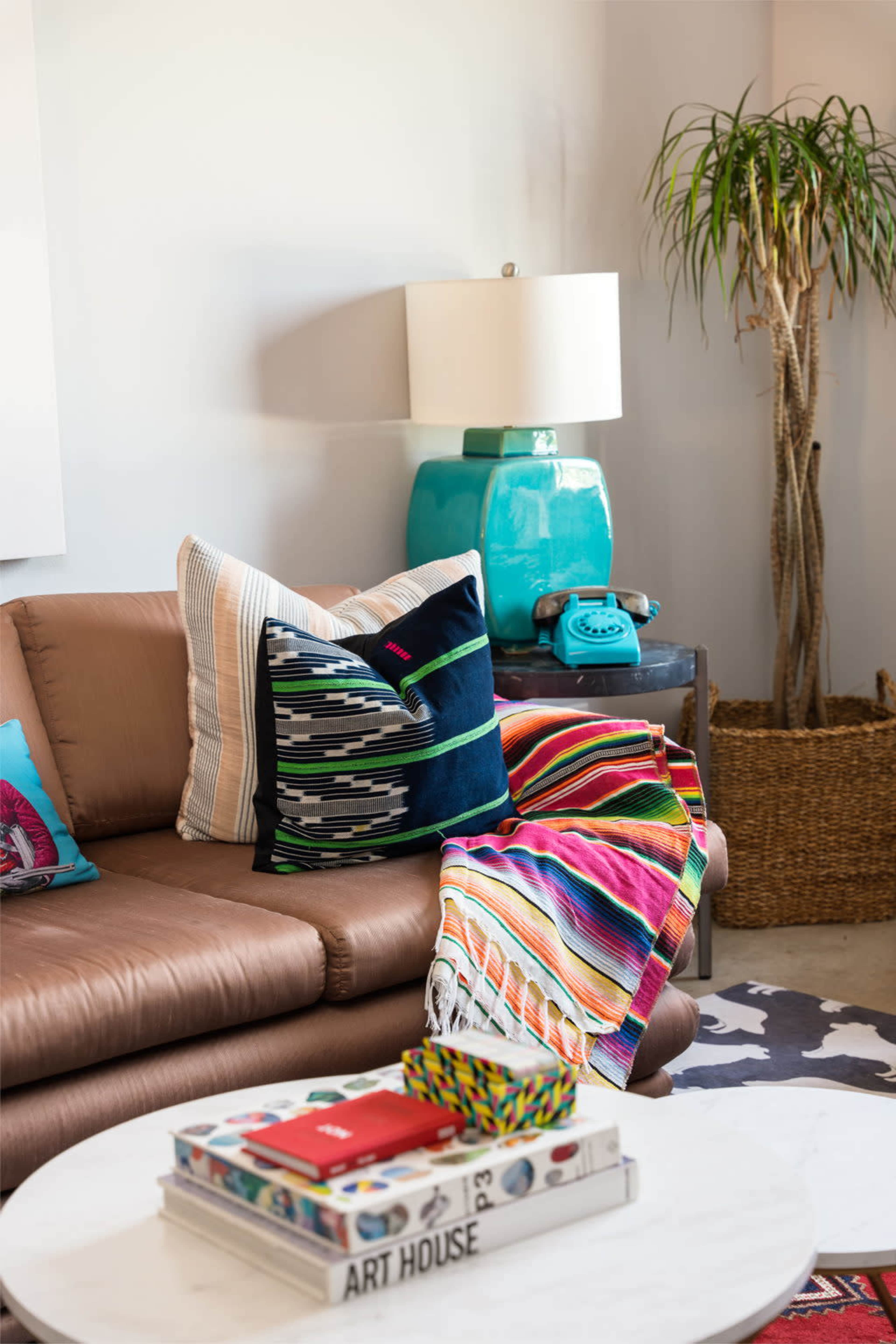 A brown sofa is adorned with colorful pillows and a striped blanket, next to a turquoise lamp and a vintage telephone on a side table.