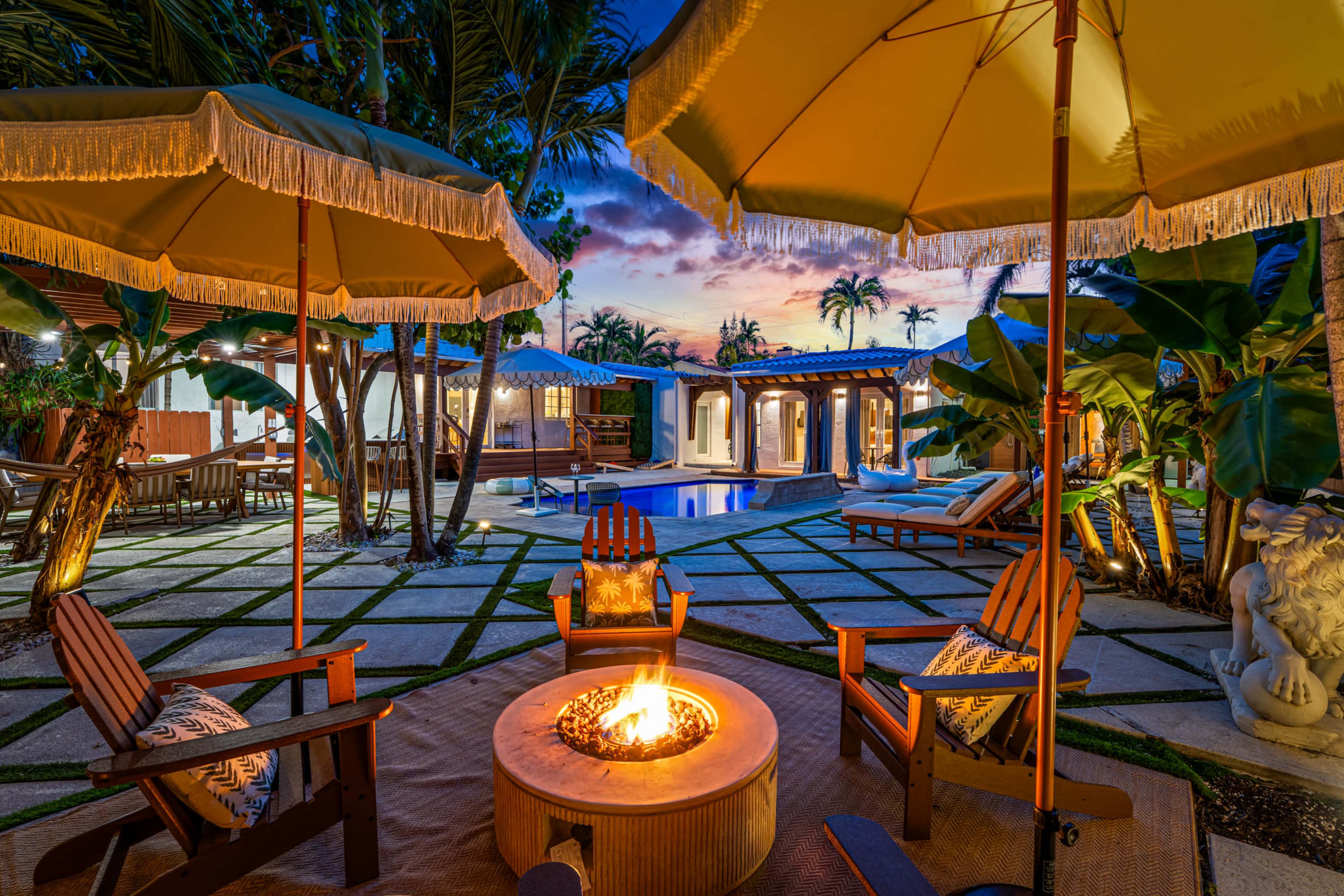 The image shows a backyard setting with two wooden chairs around a fire pit, surrounded by palm trees, umbrellas, and a swimming pool in the background at sunset.