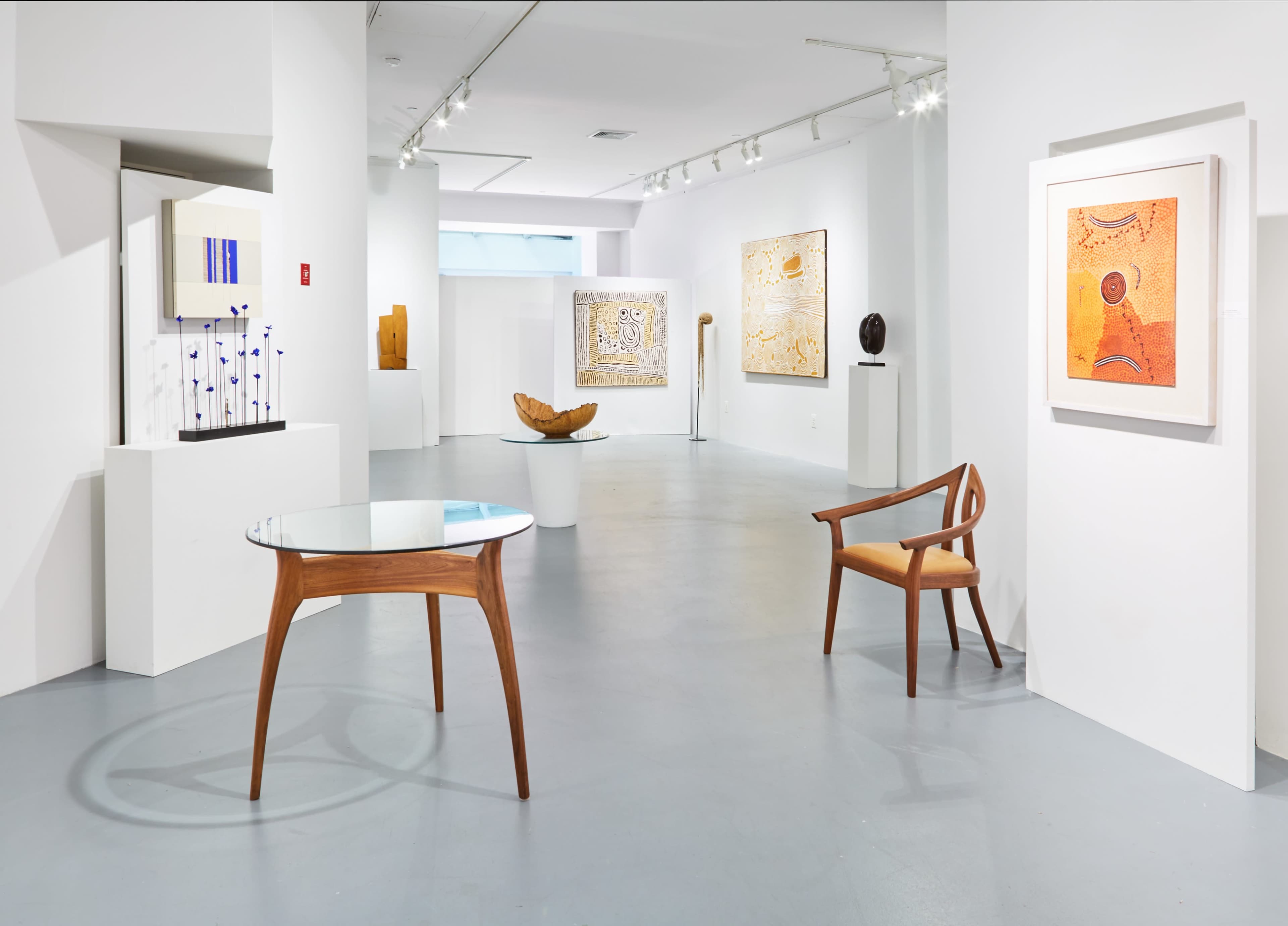 The image shows an art gallery featuring various sculptures and paintings displayed on white walls, along with a wooden table and chair in the foreground.