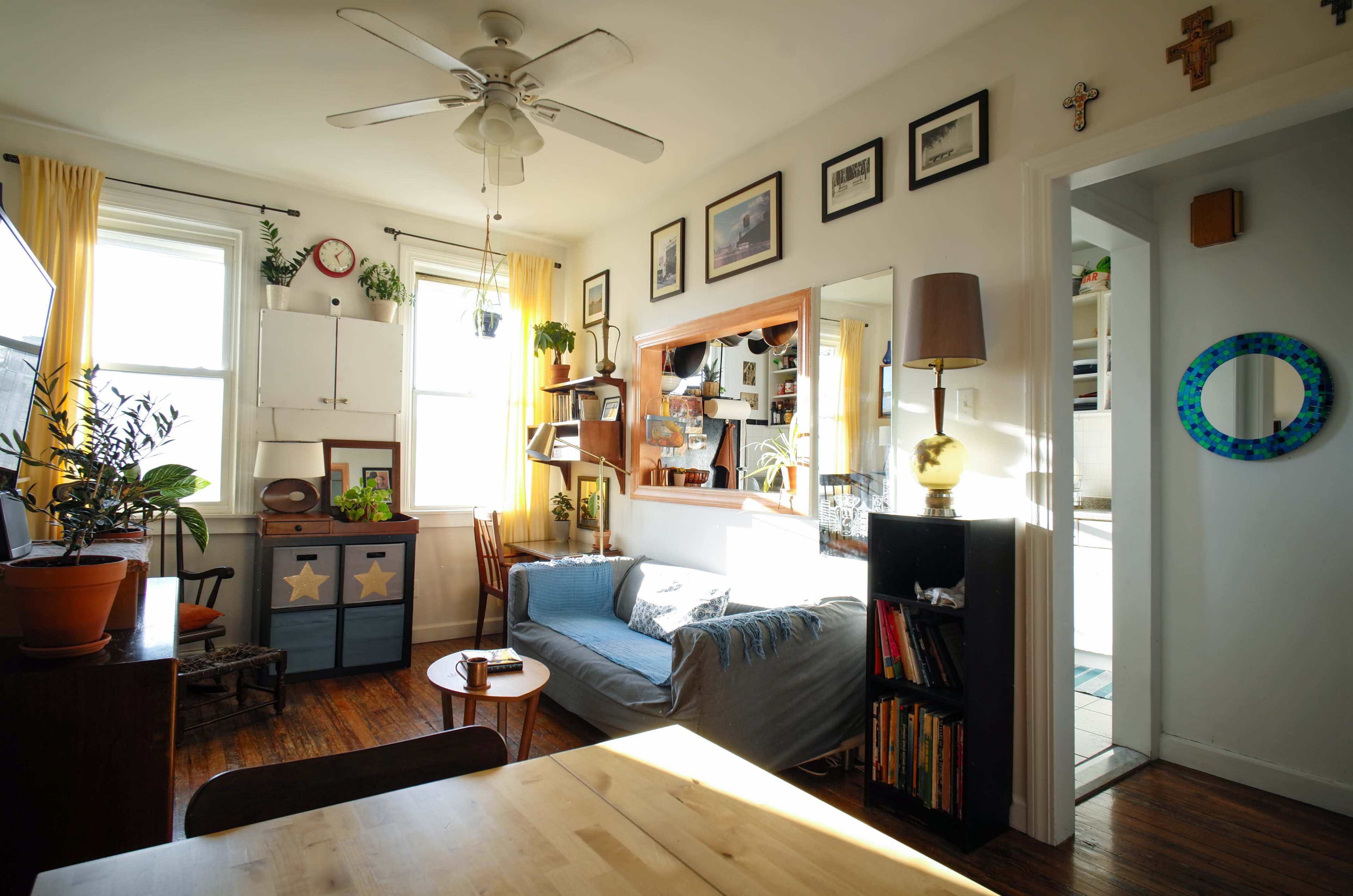 A bright living room features a blue sofa, potted plants, and various framed pictures on the walls, with a view of a nearby kitchen through an open doorway.