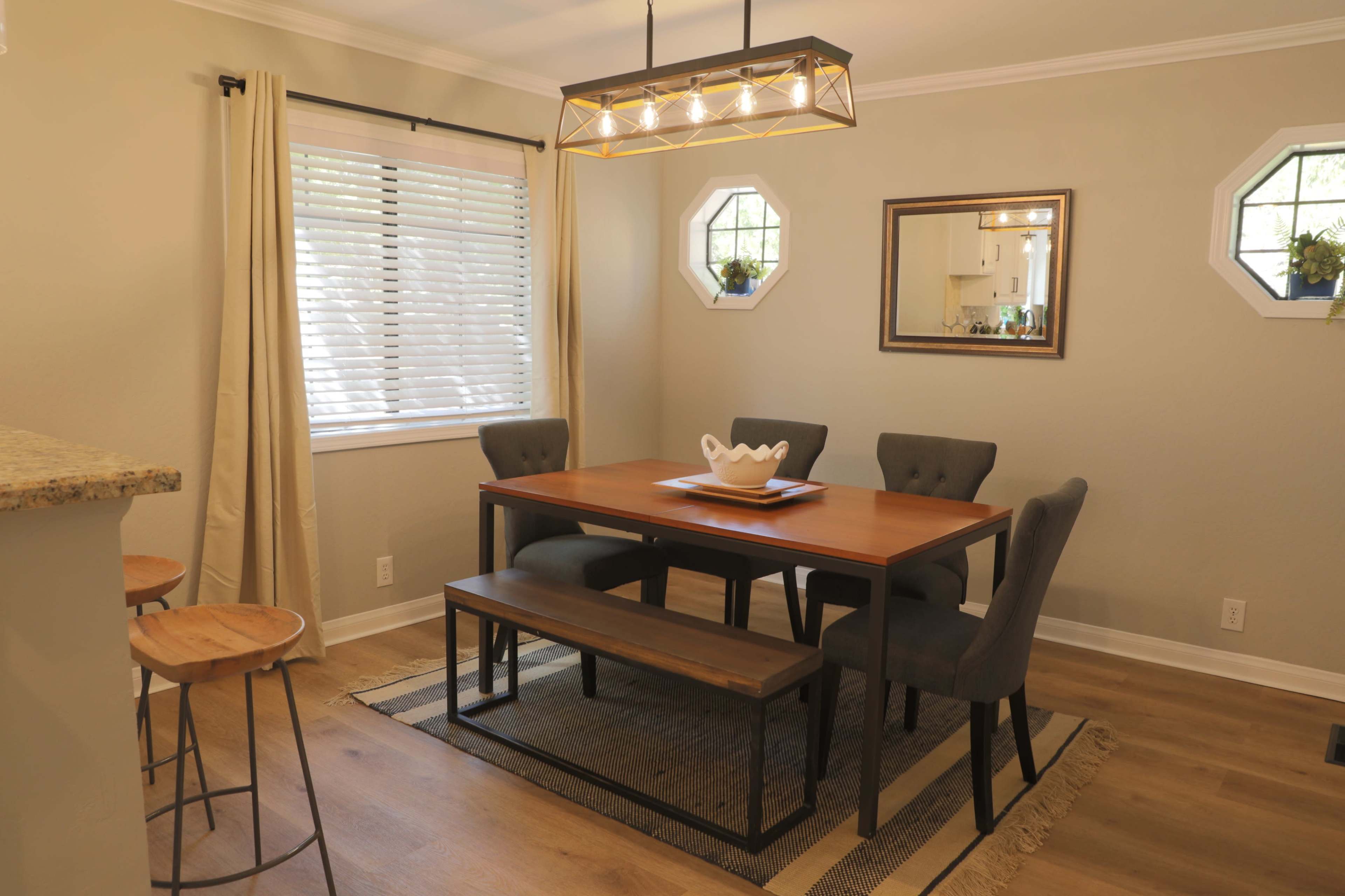 A dining area features a wooden table with upholstered chairs and a bench, illuminated by a modern chandelier and surrounded by windows.