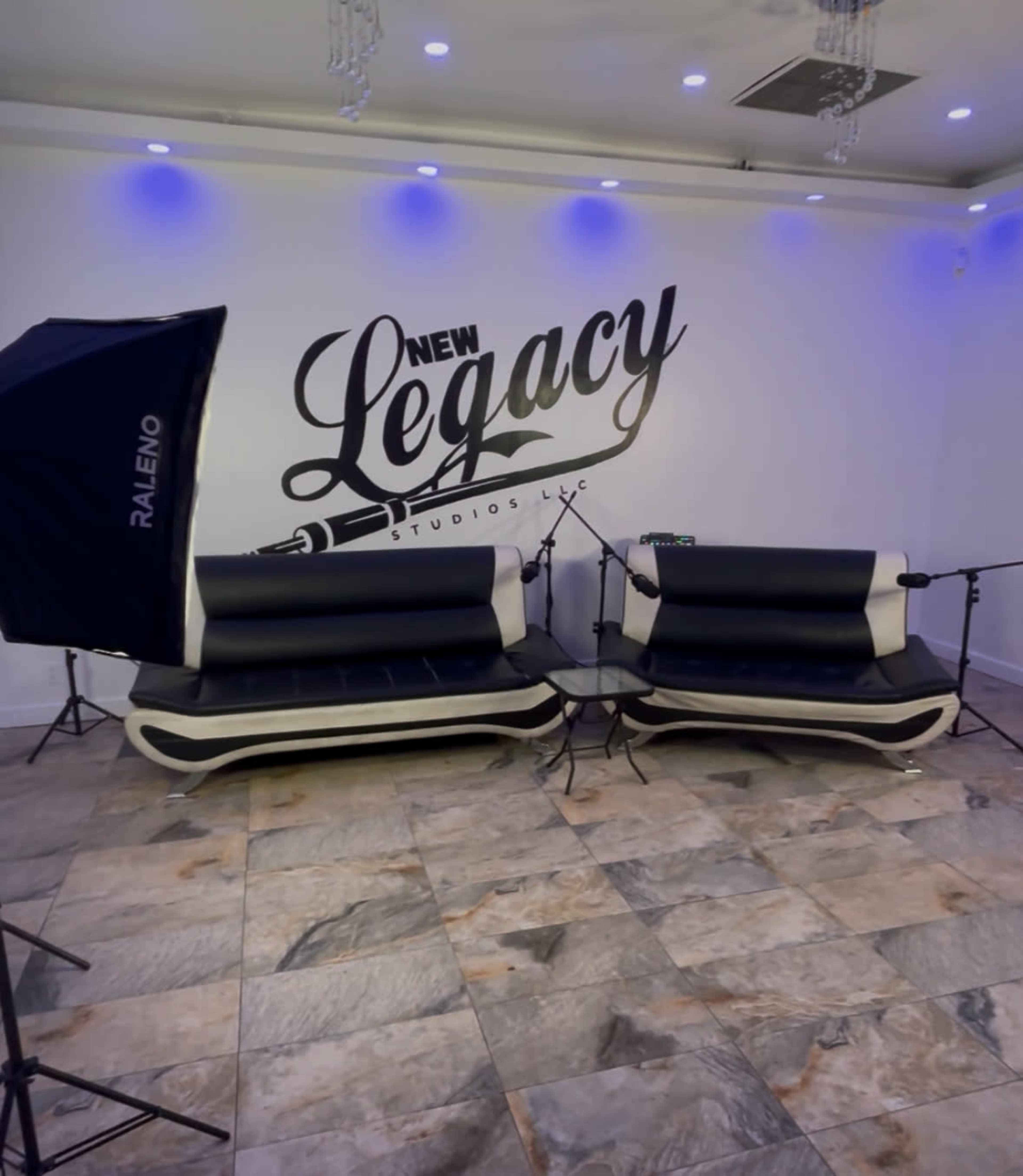 The image shows a modern recording studio with two black and white couches and microphones set up against a wall featuring the phrase "New Legacy Studios LLC."