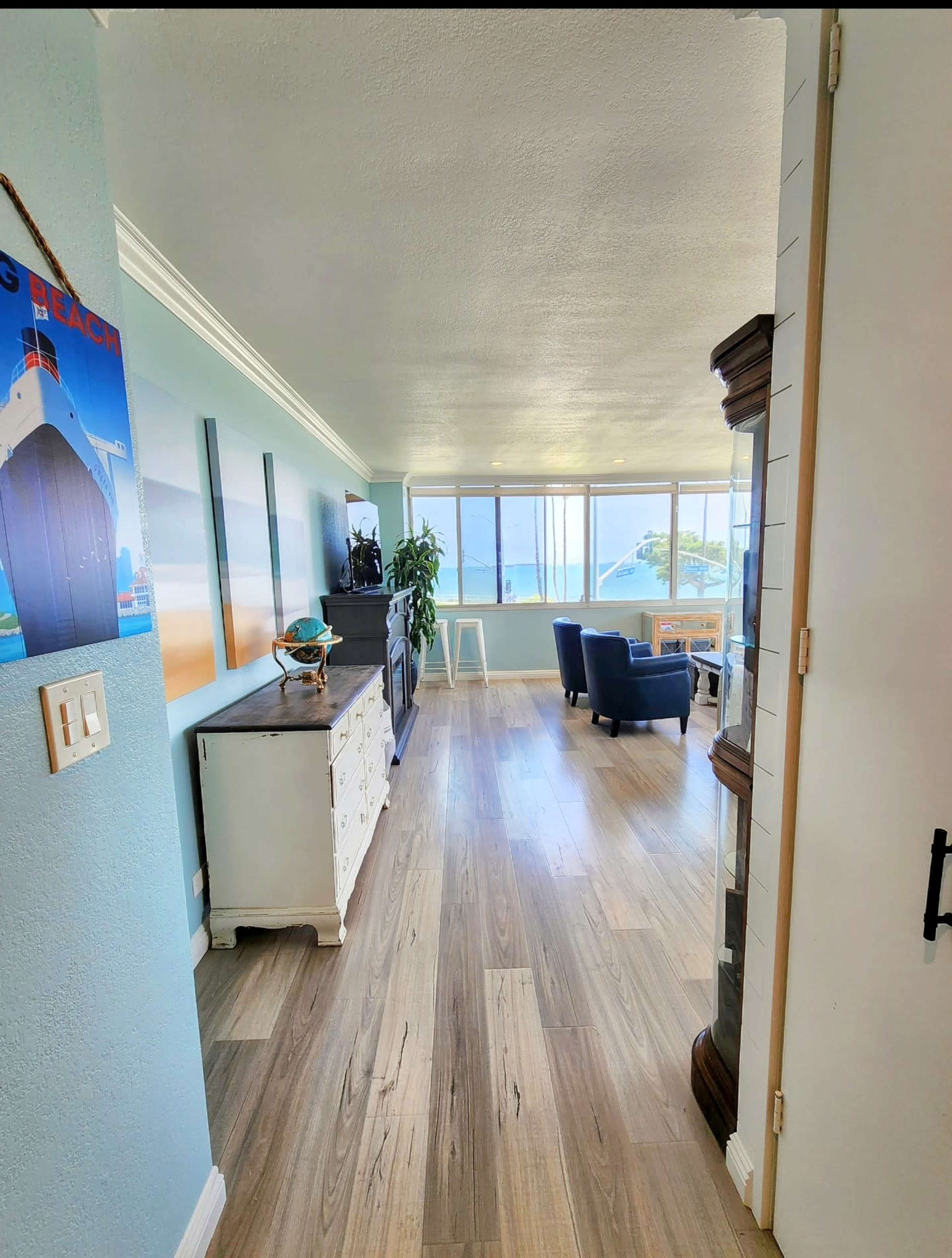 A bright interior space with wood-style flooring, a living area featuring blue chairs, and large windows that overlook a view outside.