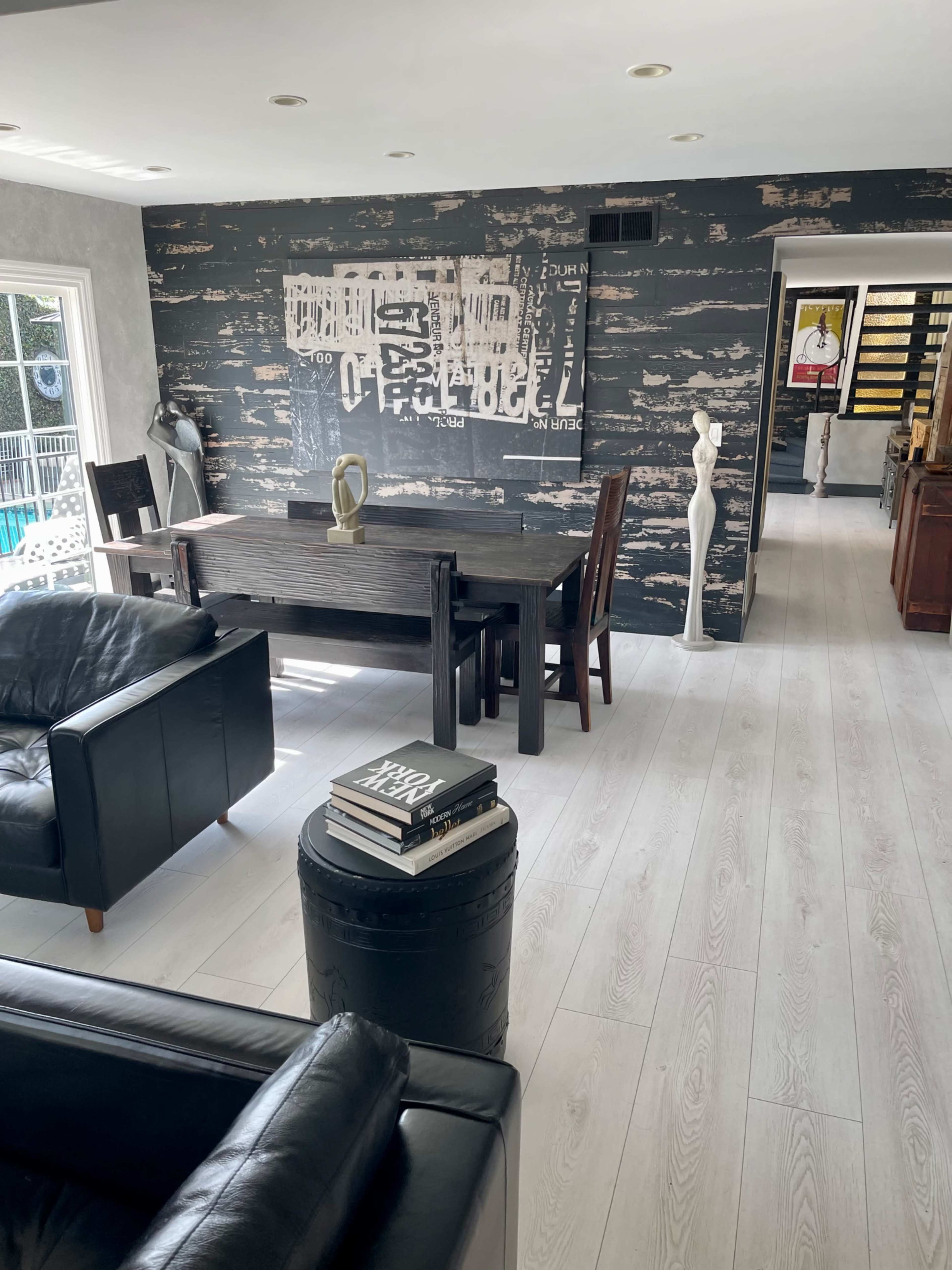 The image shows a modern living space with a dark wooden dining table, a black leather sofa, and a textured wall featuring an abstract artwork.