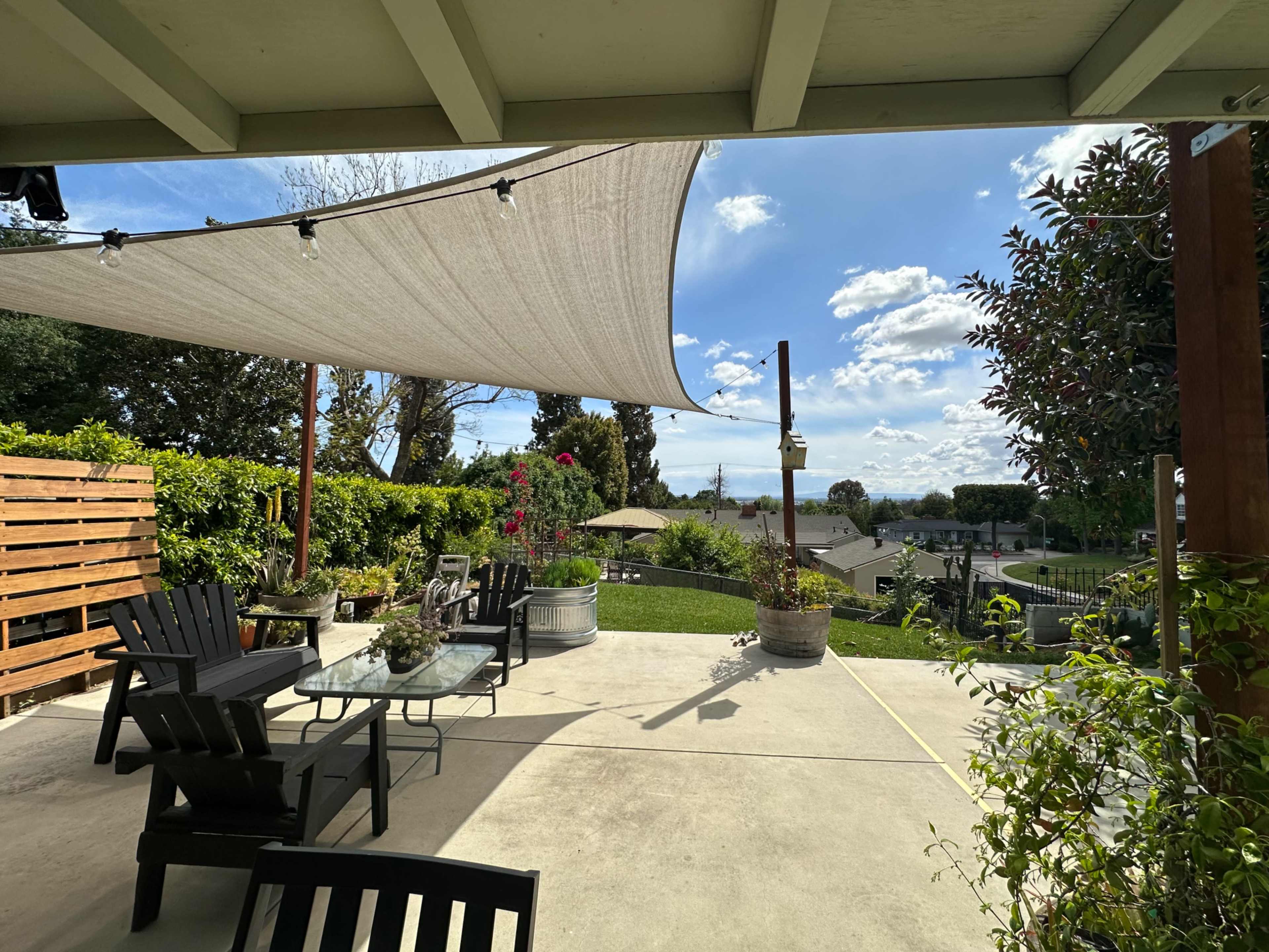 A covered patio features black outdoor chairs, potted plants, and a view of a green lawn and distant hills under a blue sky with scattered clouds.