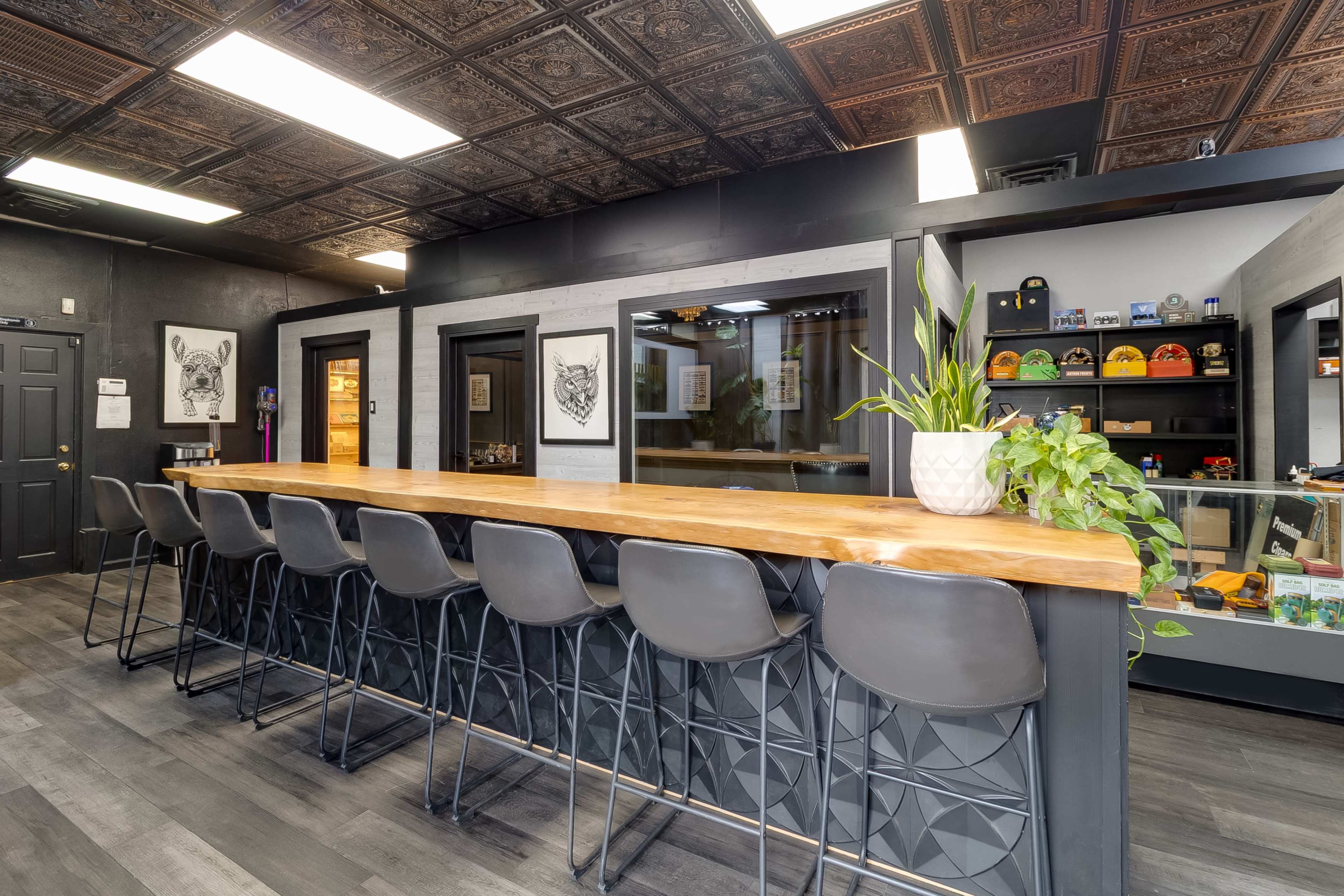 The image shows a modern interior featuring a long wooden bar with high black stools, framed by dark walls and decorative ceiling tiles.
