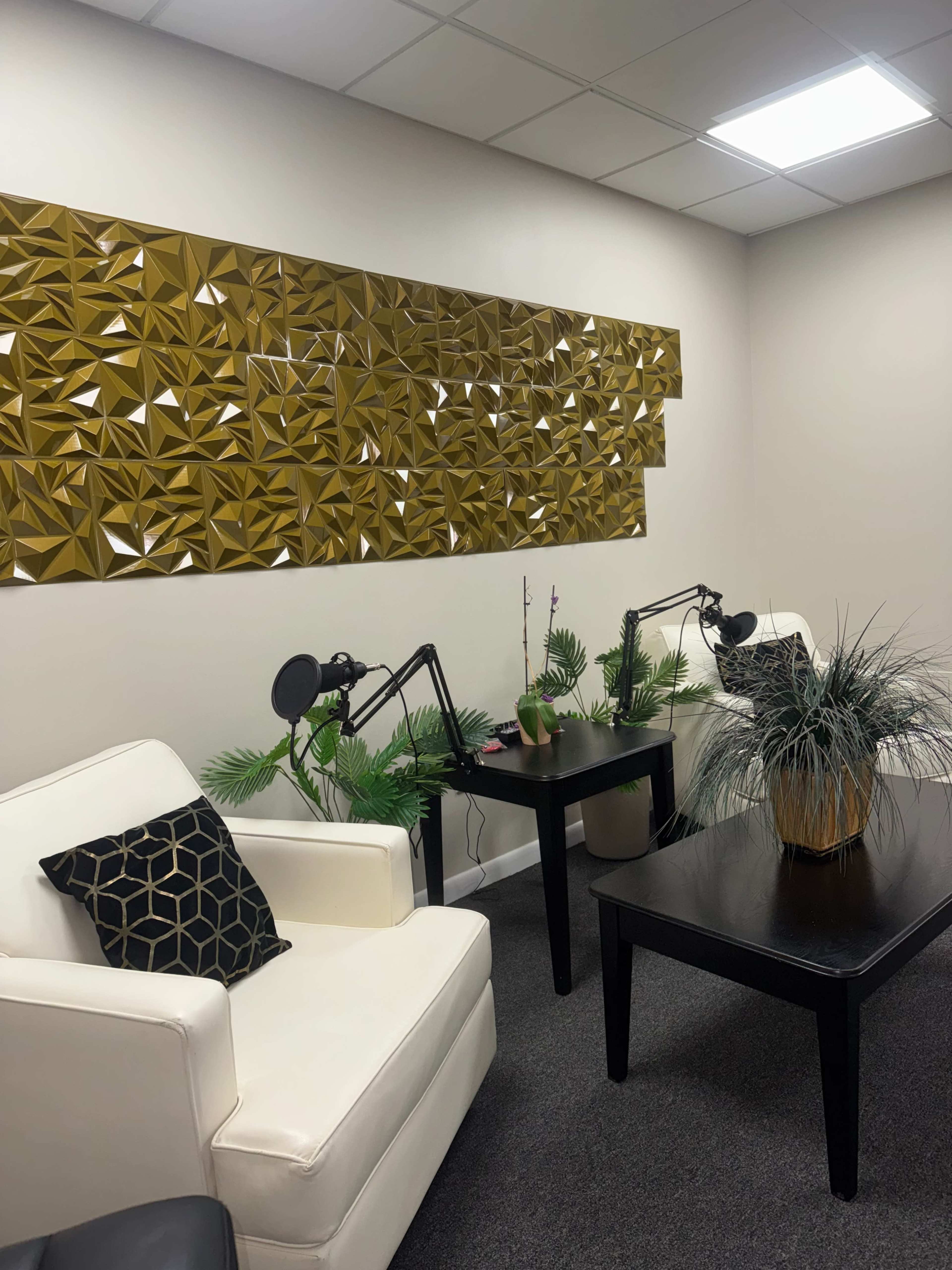 The image shows a small, modern office space featuring two white armchairs, a dark wooden table, and a decorative gold wall panel.