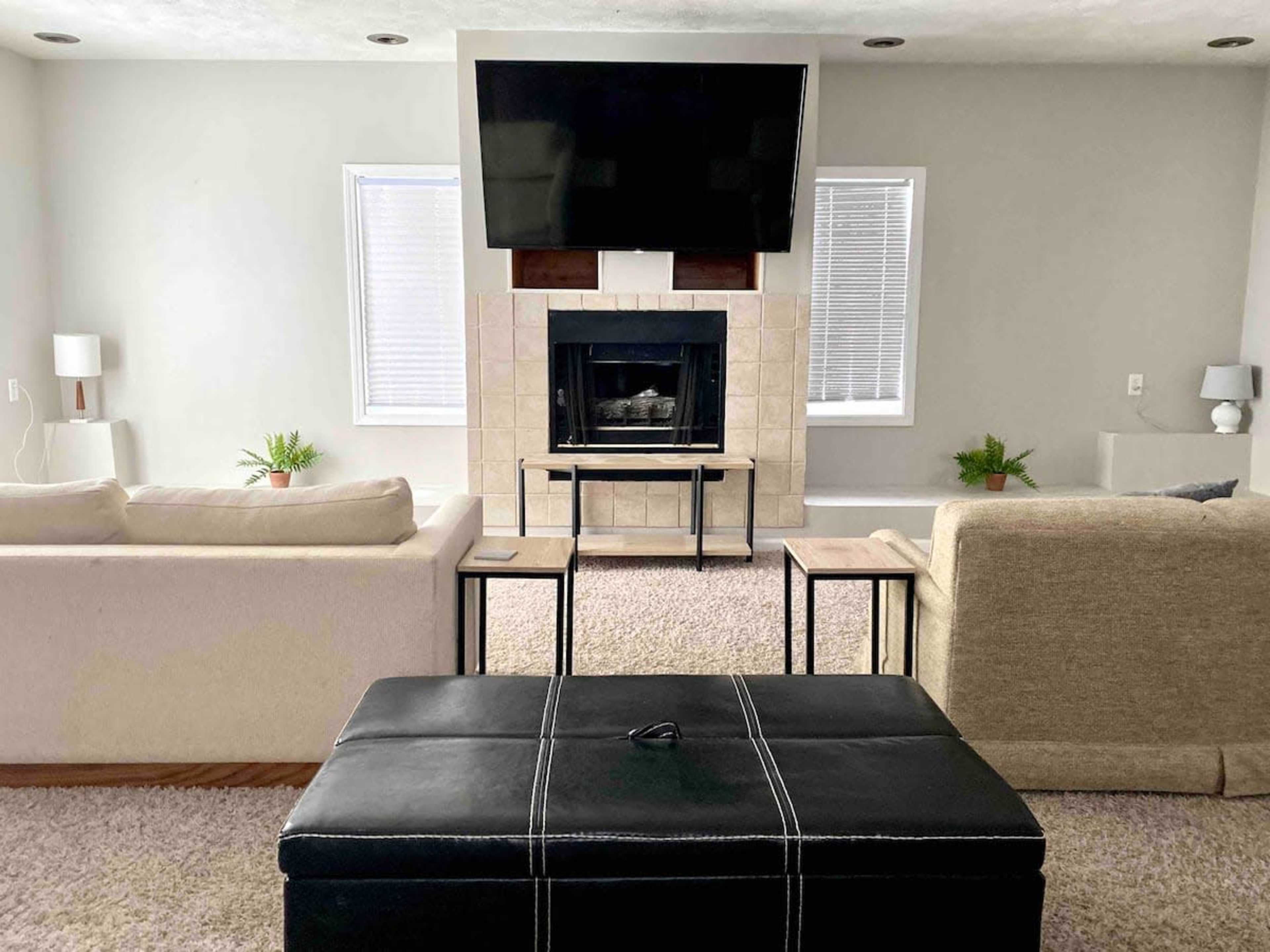 A modern living room with a fireplace, a large television mounted above it, and two sofas facing each other, with a black storage ottoman in the center.