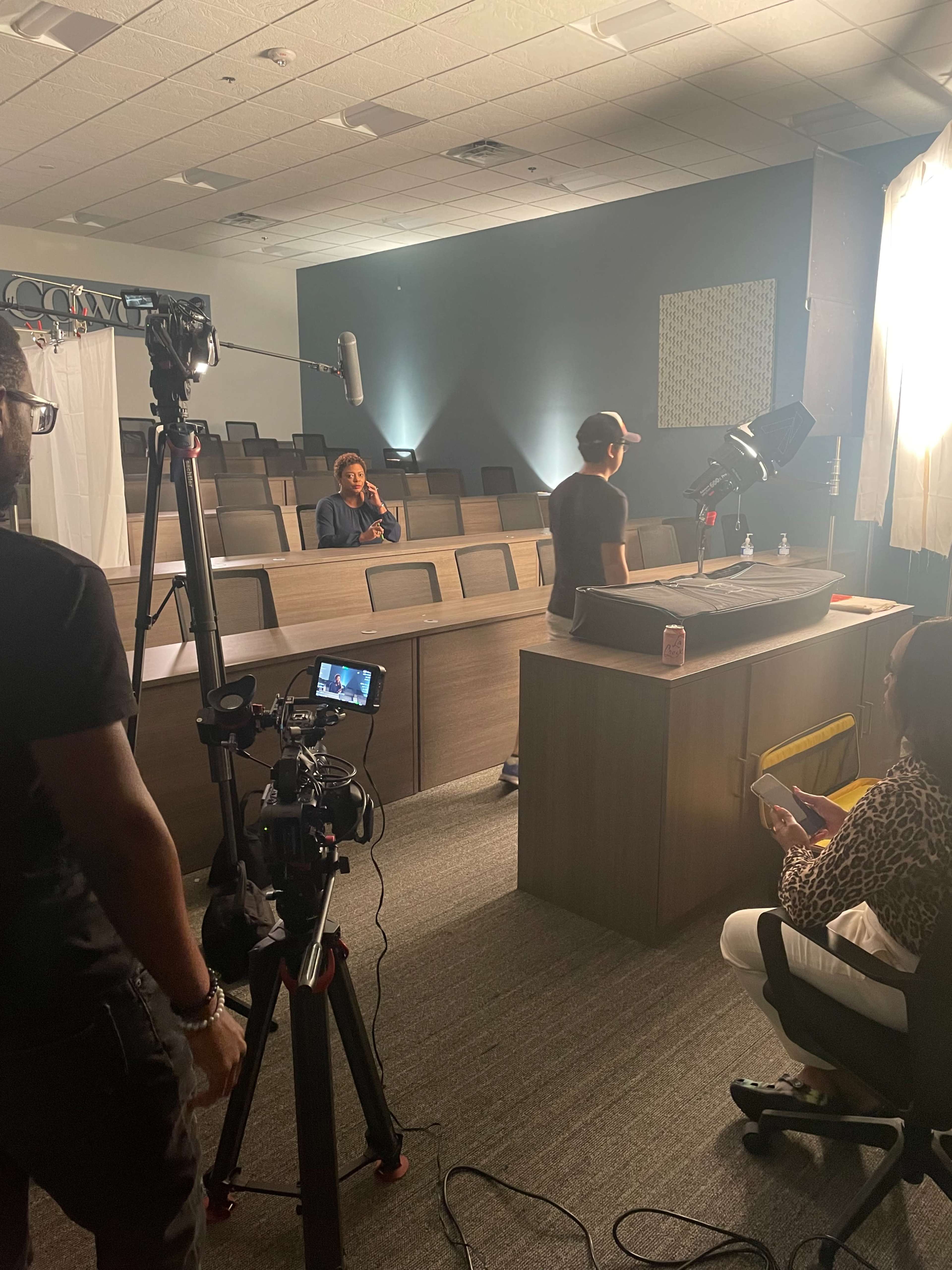 A film crew is setting up equipment in a classroom for a video shoot, with a few people positioned near a desk.