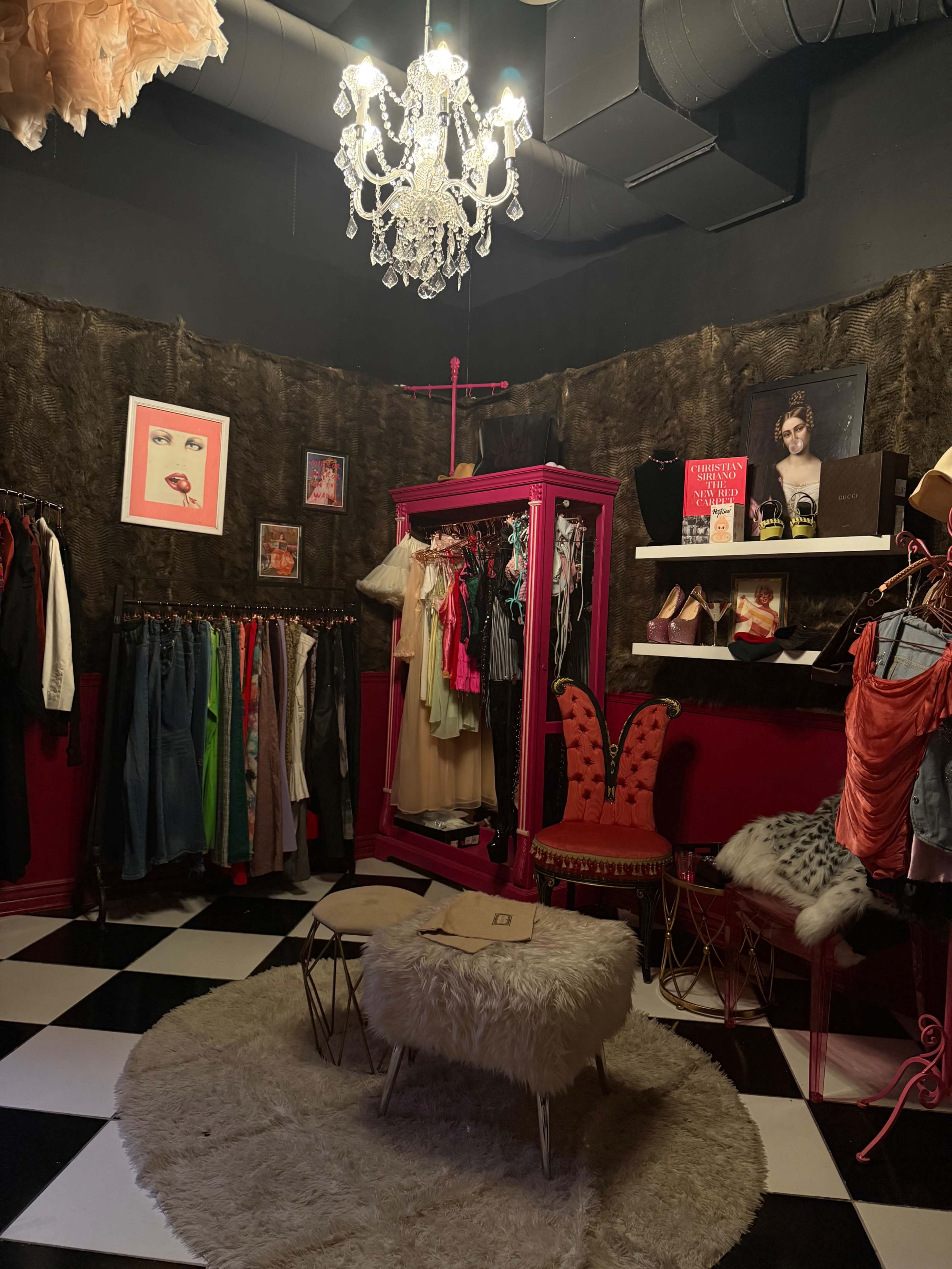 The image shows a boutique-style dressing room with a pink garment rack, various clothing items hung on walls, and a plush seating area featuring a furry ottoman.
