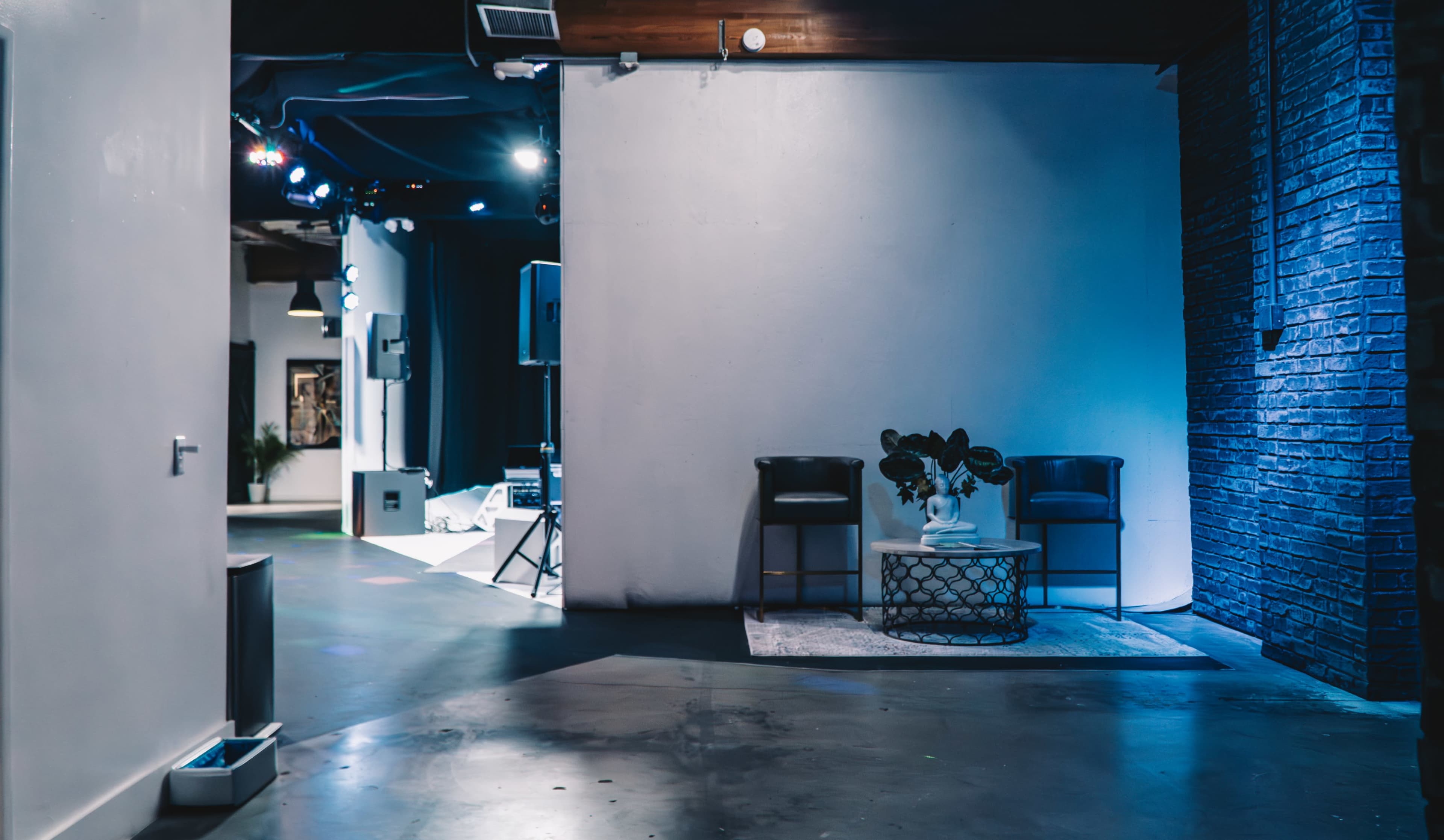 A modern event space with two chairs and a table in the foreground, and a stage area with lights in the background.